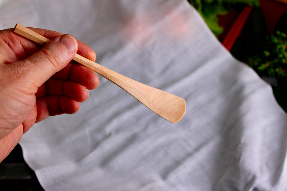 hand holding a long thin wooden tan jam spoon showing wood grain on white cloth background with kitchenware
