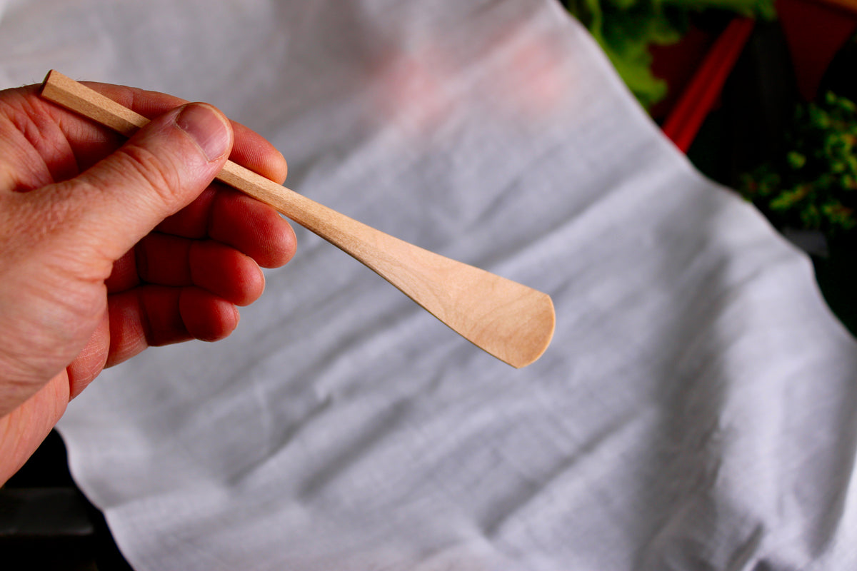 hand holding a long thin wooden tan jam spoon showing wood grain on white cloth background with kitchenware