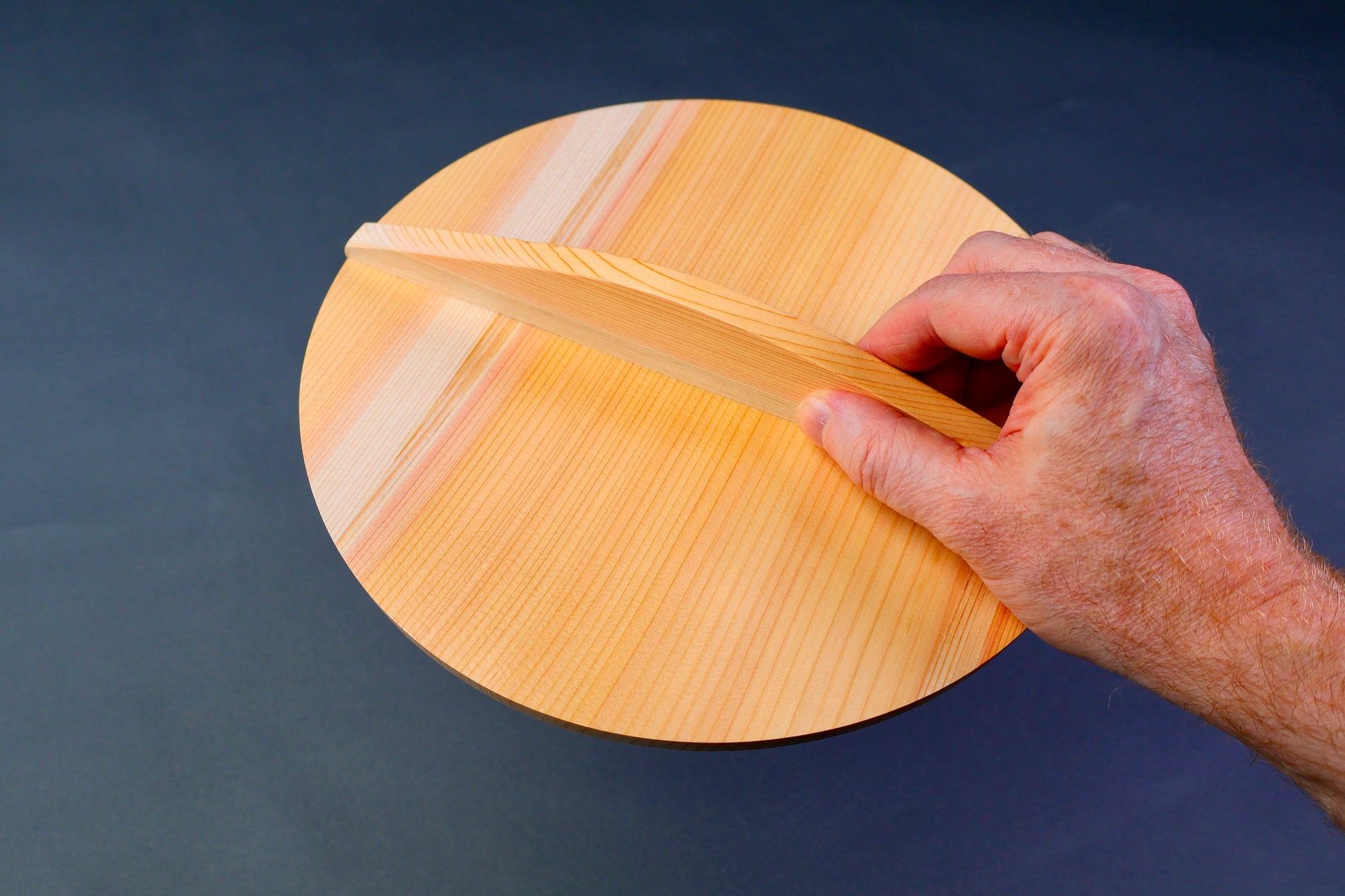 hand holding circular wooden drop lid with long handle in grayish blue backdrop