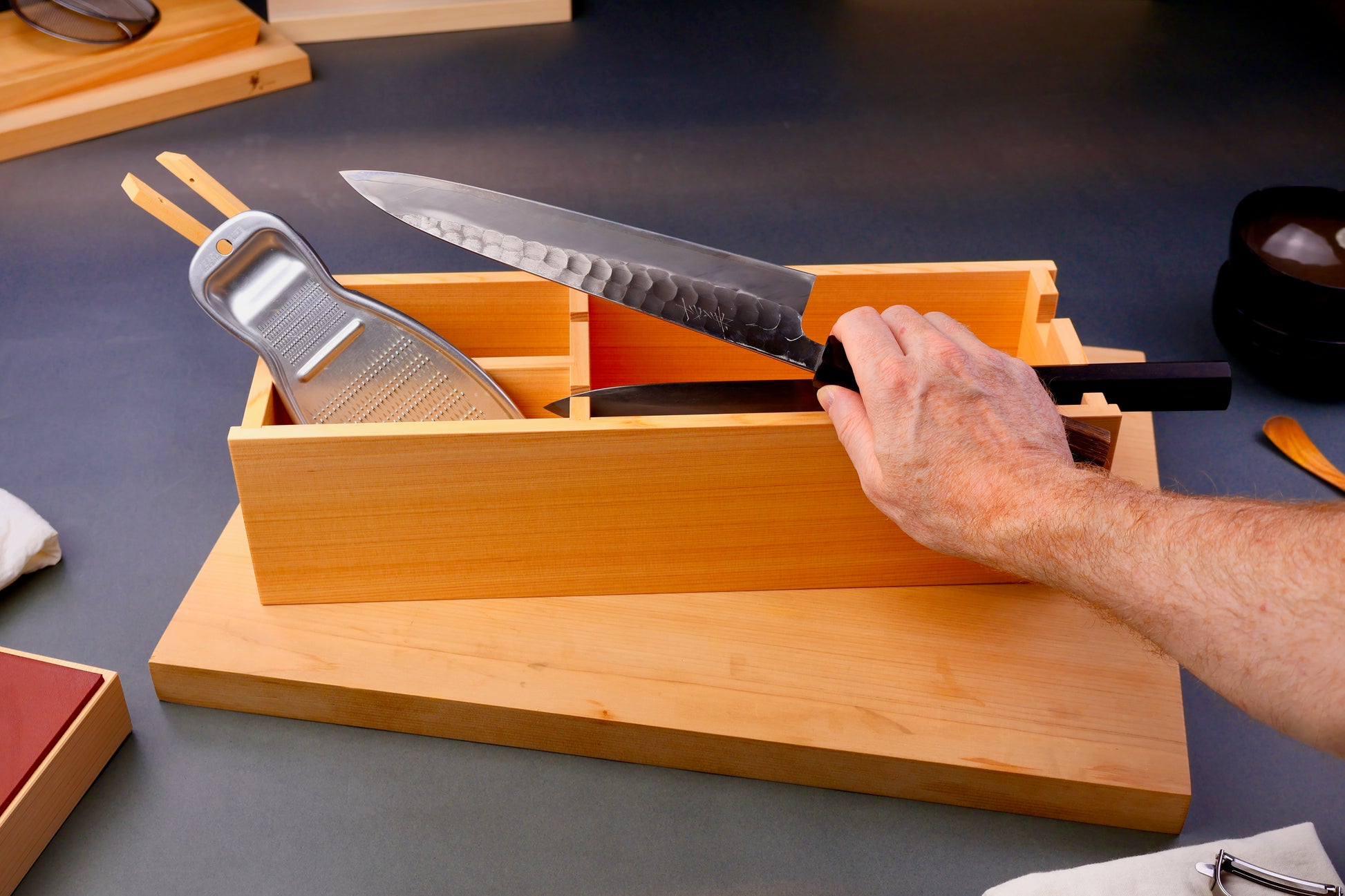 hinoki wood rectangular shaped box with natural wood grain containing knives and tools sits atop wood cutting board and hand holding knife surrounding japanese kitchenware on gray background