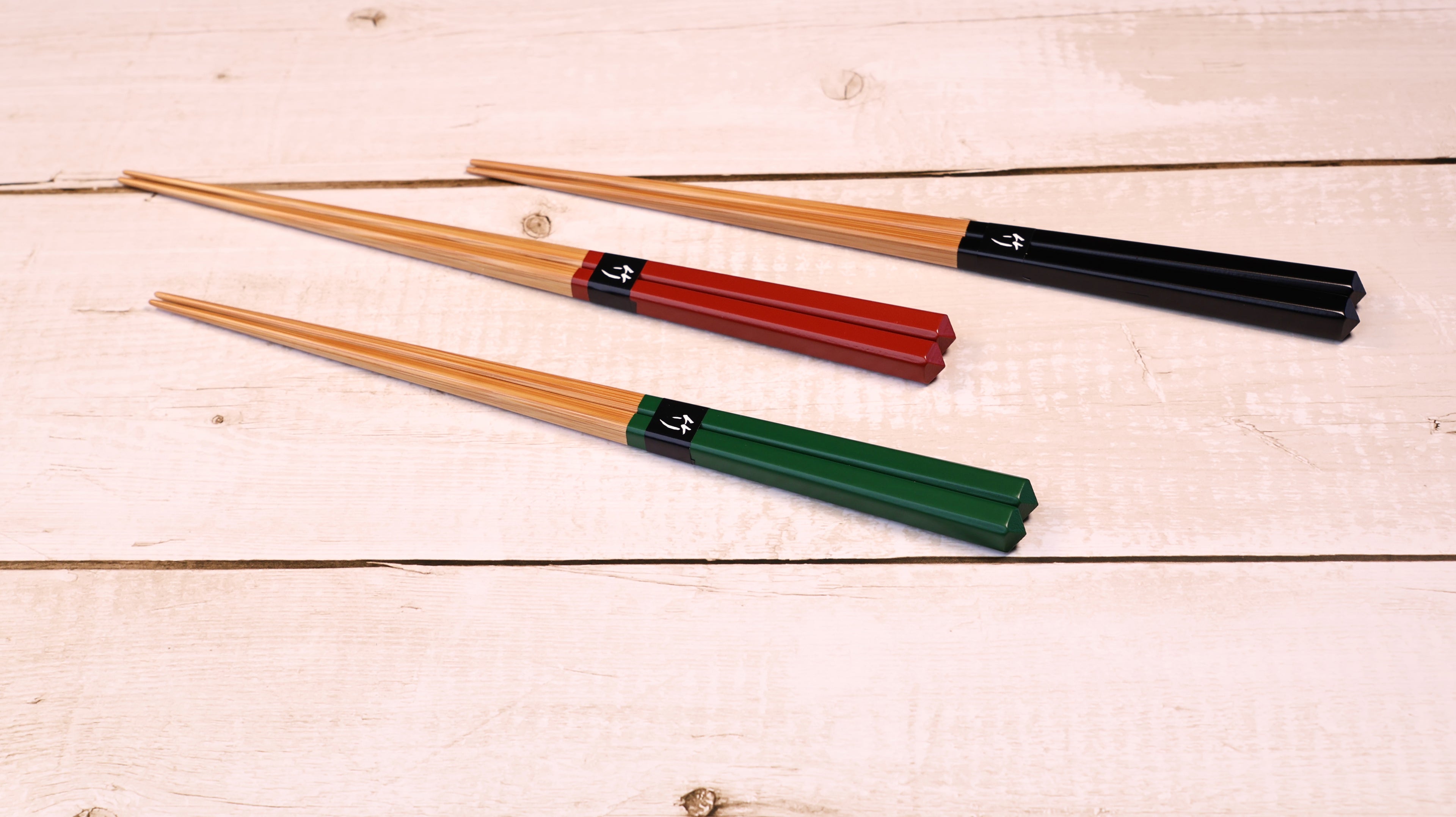 Three pairs of wooden chopsticks with colored handles in green, red, and black, arranged diagonally on a light-colored wooden plank surface.