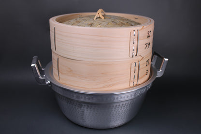 detailed shot of round handmade hinoki steamer with lid and base sitting atop steam ring and aluminum two handled steaming pot the grey backdrop is in contrast to prominent image of products