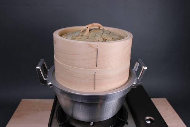 chinese steamer artisan handmade in japan extra thick body sits atop aluminum steam ring and dantsuki seiro steam pot made of aluminum atop a portable gas stove and hinoki cutting board and a grey walled background