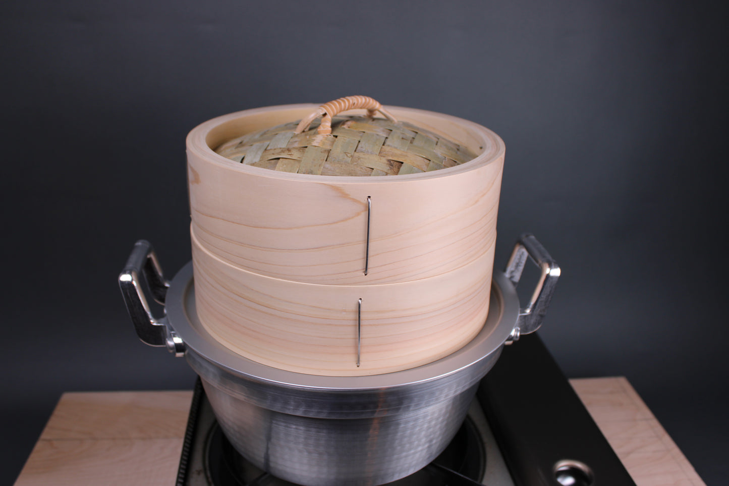 chinese steamer artisan handmade in japan extra thick body sits atop aluminum steam ring and dantsuki seiro steam pot made of aluminum atop a portable gas stove and hinoki cutting board and a grey walled background