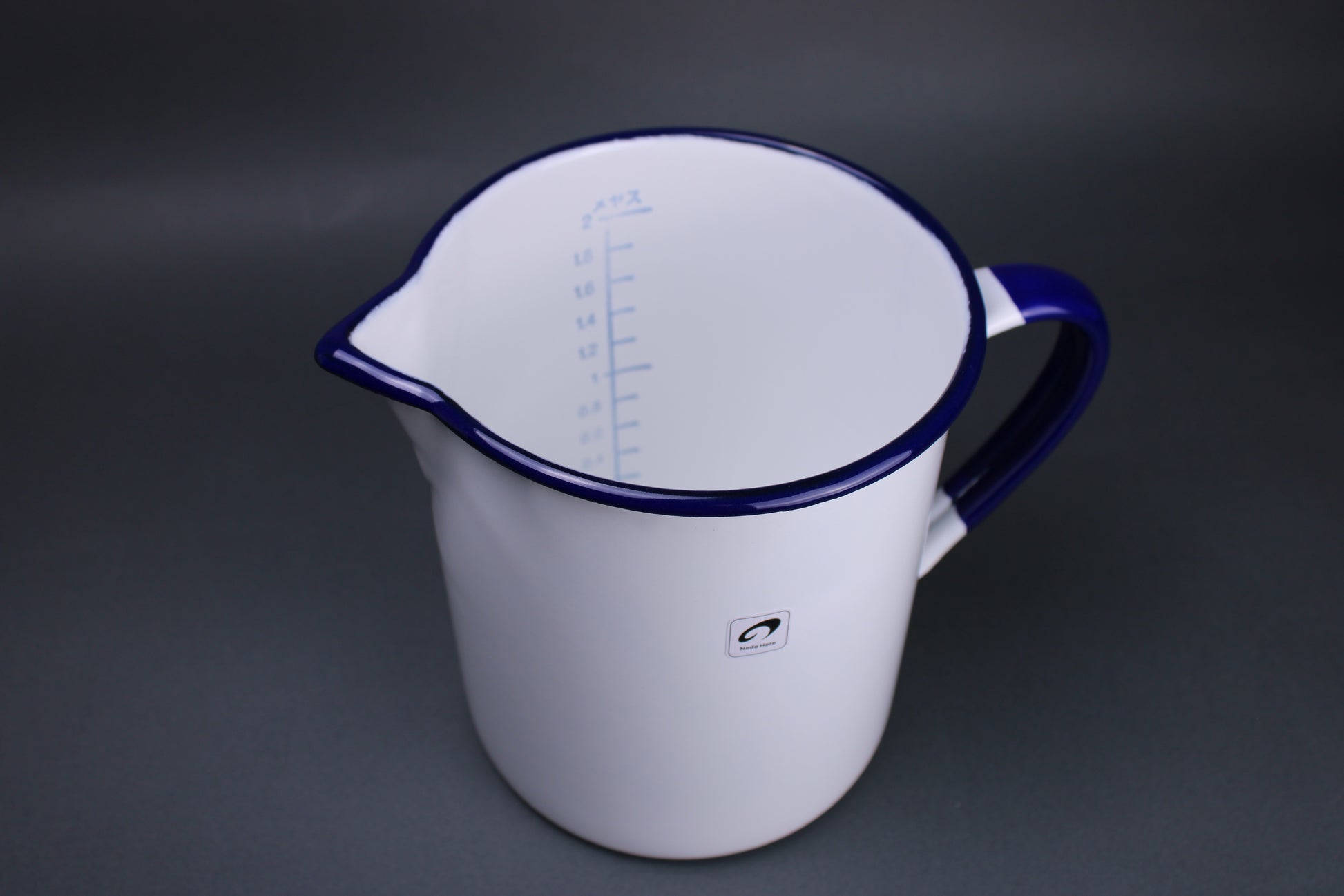 top down view of a two liter measuring cup featuring the metric measurements  printed inside the cup and node horos signature white glazed body and navy blue trimmed handle and rim there is a grey background to contrast the product