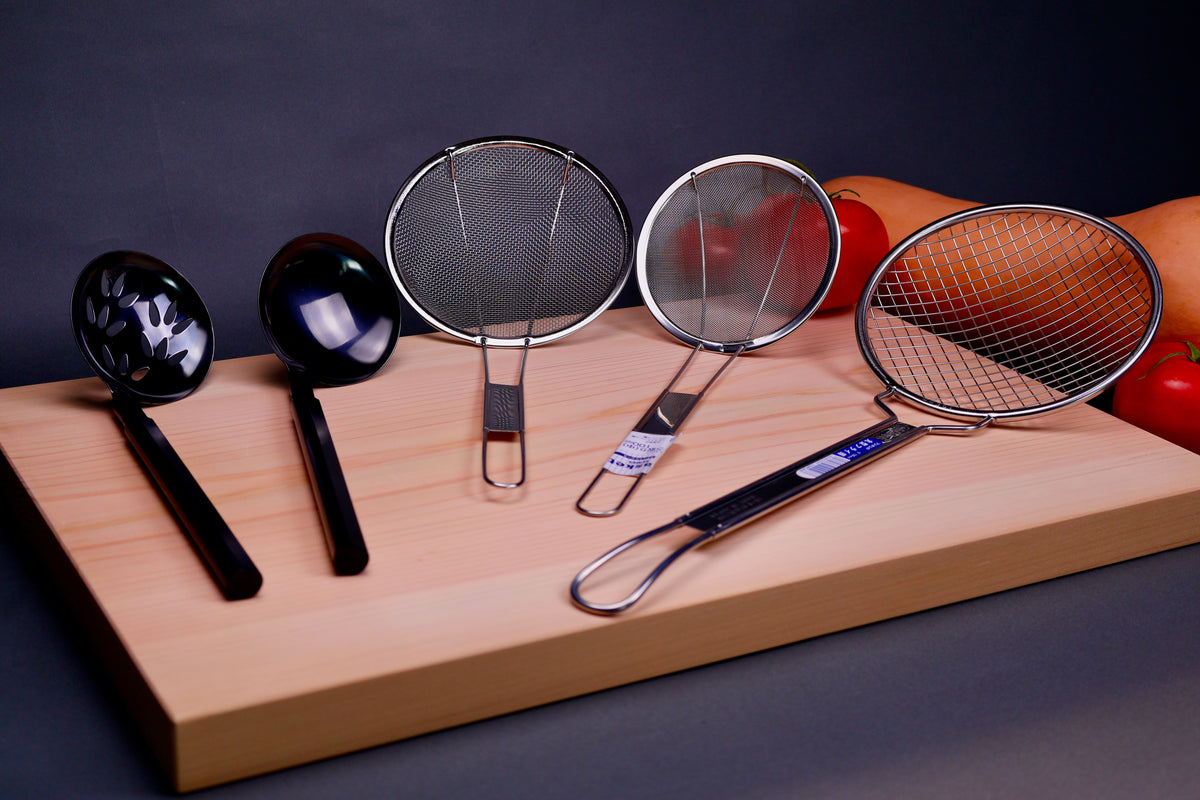 two kitchenware serving ladles and three fine to coarse mesh skimmers lay atop hinoki cutting board beside tomatoes and squash in grey backdrop 