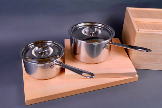 two shiny art deco steel saucepans with curved lids on wood cutting boards beside wood rice box in light grey back ground