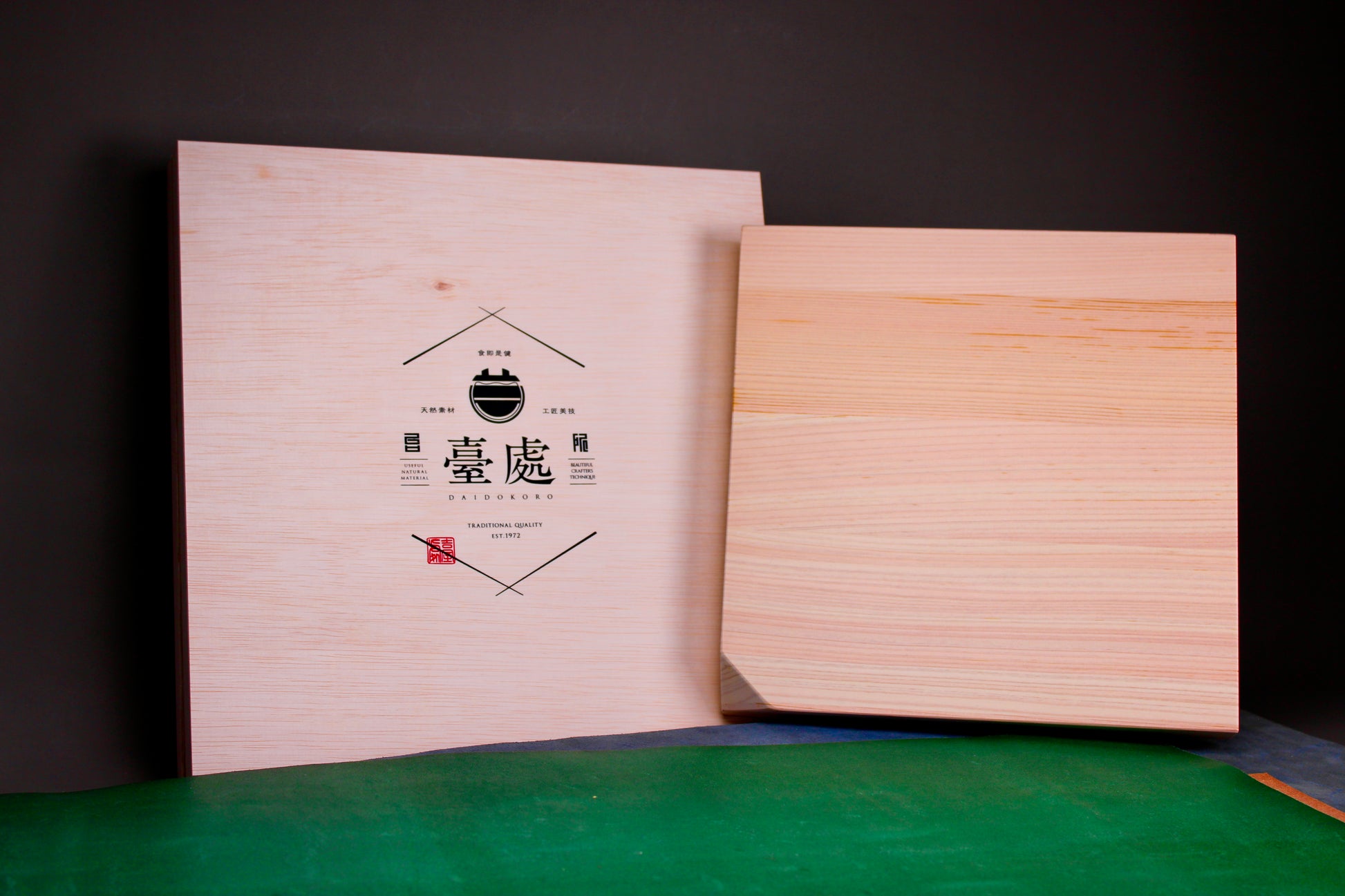 square shaped lightly tanned kiso hinoki cutting board beside tan whitish paulownia wood gift box with hiragana print on green and blue surface in black backdrop