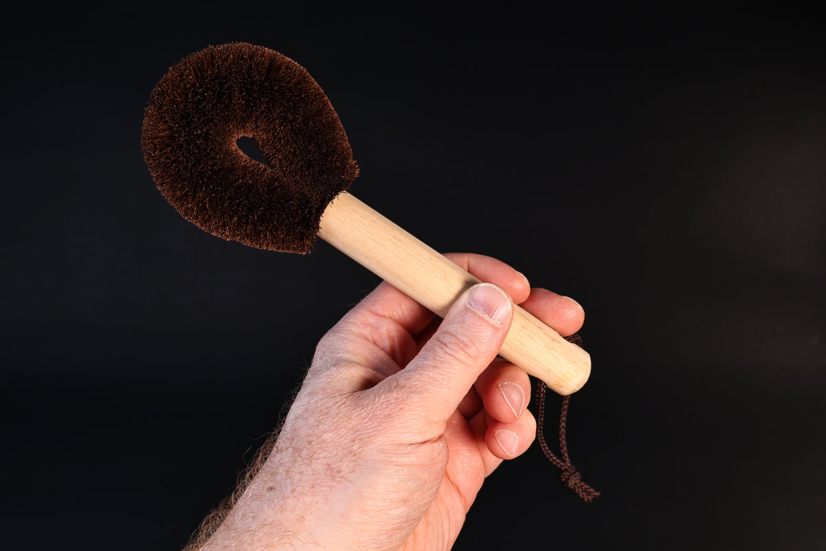 Kitayama Masatsumu tawashi brush held up by the wooden handle, showing the rounded palm-fiber head and cord loop