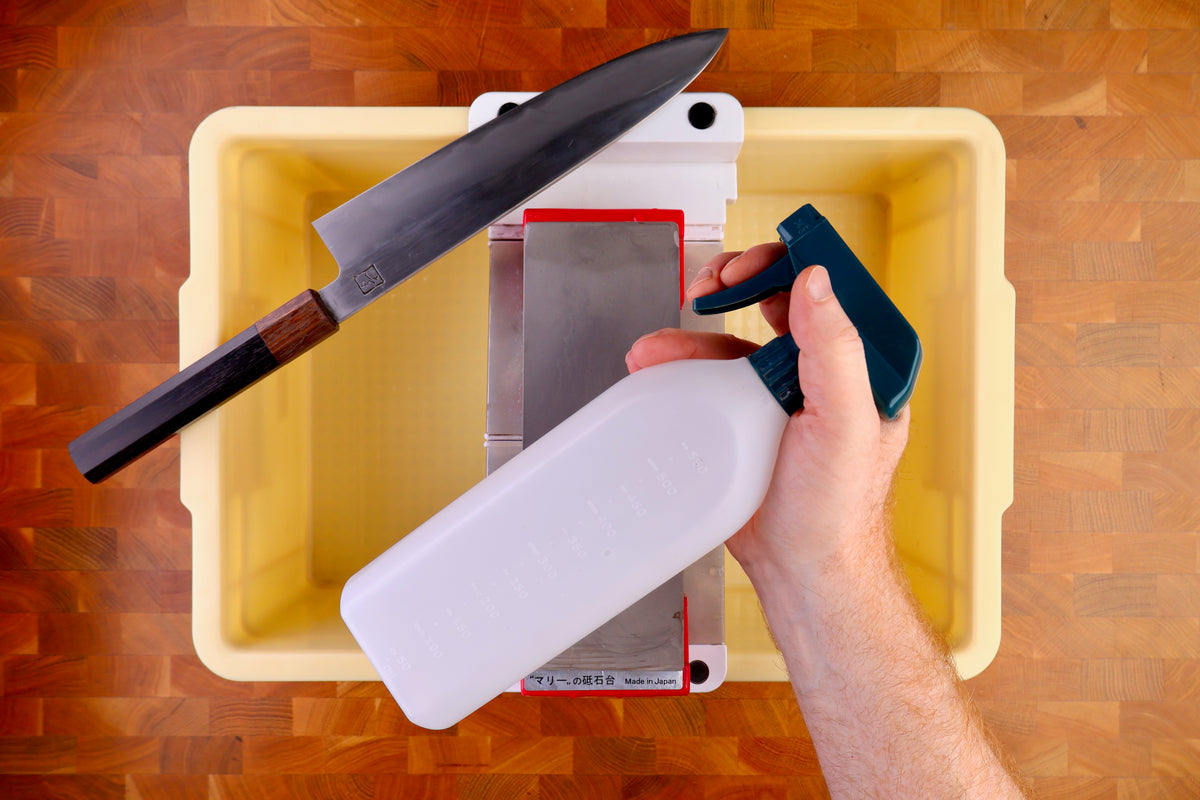 tall yellow deep plastic box holding sharpening bridge whetstone hand holding plastic spray bottle wood handled knife atop wood countertop 