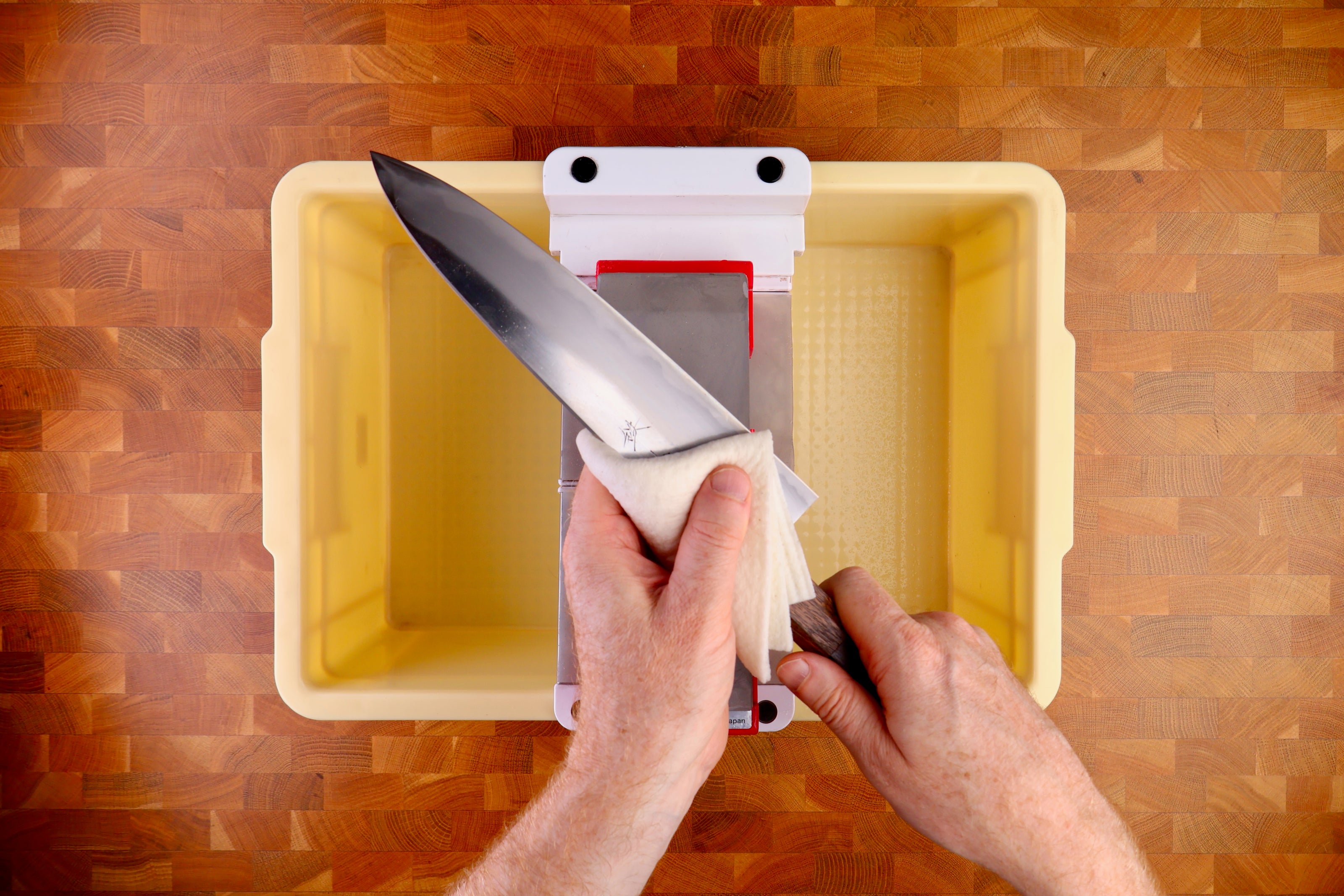 tall yellow deep plastic box holding sharpening bridge whetstone deep white cloth being wiped by hand on wood handled knife atop wood countertop 