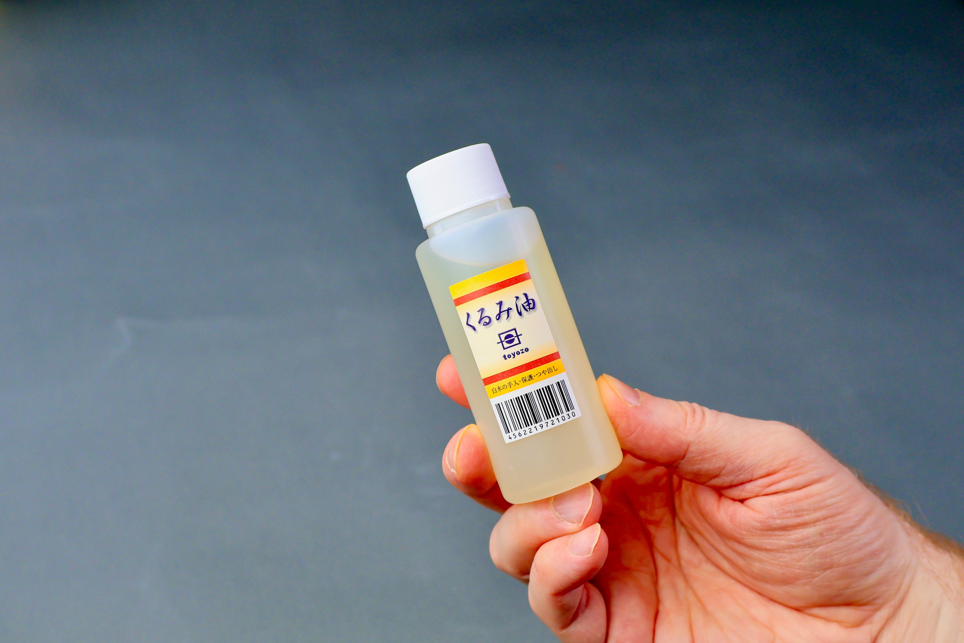 hand holding a small clear plastic bottle with yellow red stripe Toyoza brand with ups code in grey backdrop