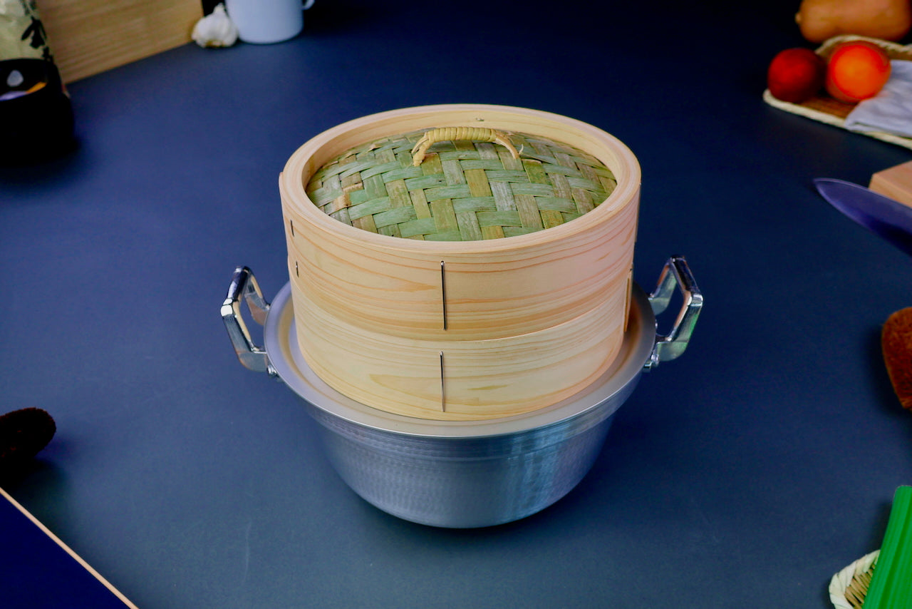 thick wood round bamboo steamer set woven top with loop natural wood tone with printed hiragana sits atop double handled metal hammered pot surrounded by japanese kitchenware in blue gray background