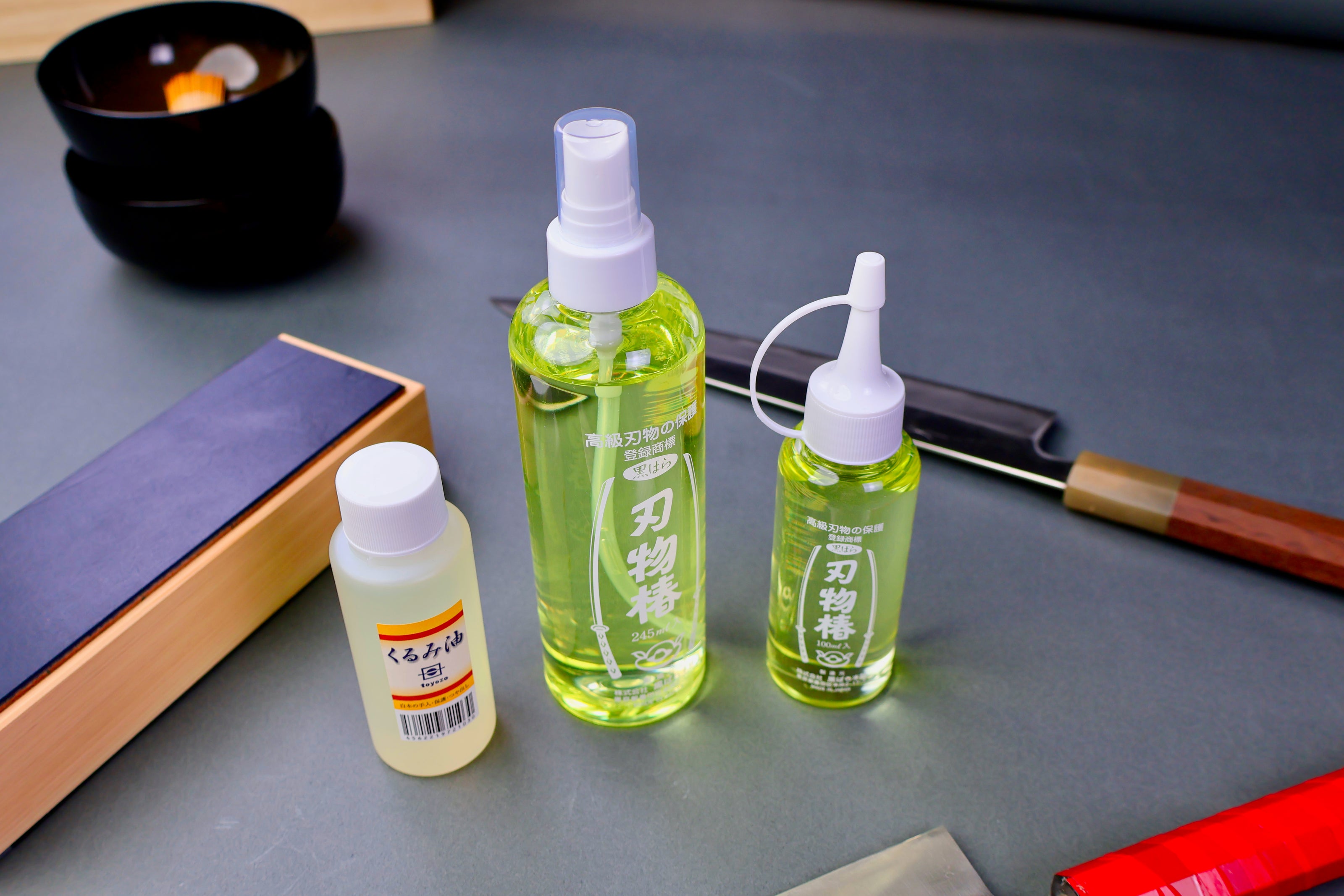 three small plastic containers for kurumi oil and Tsubaki oil surrounded by japanese knife accessories in a grey blue background