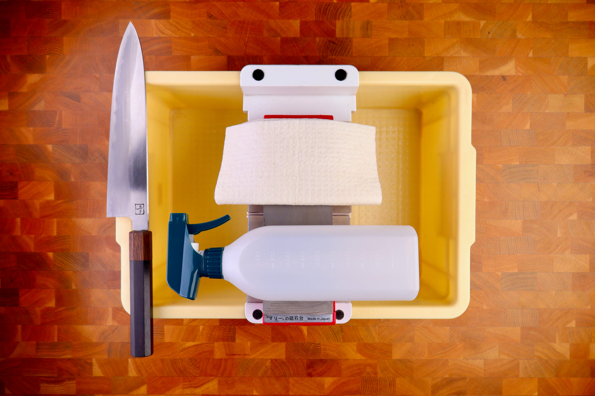 tall yellow deep plastic box holding sharpening bridge whetstone deep white cloth plastic spray bottle wood handled knife atop wood countertop 