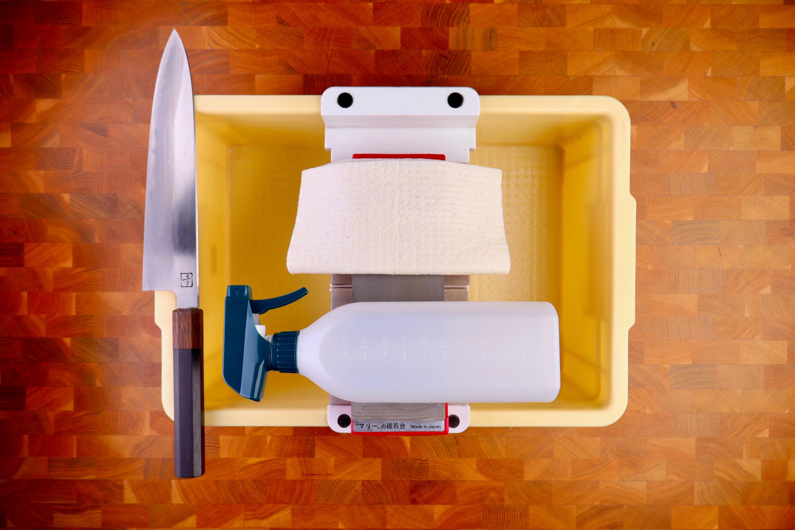 tall yellow deep plastic box holding sharpening bridge whetstone deep white cloth plastic spray bottle wood handled knife atop wood countertop 