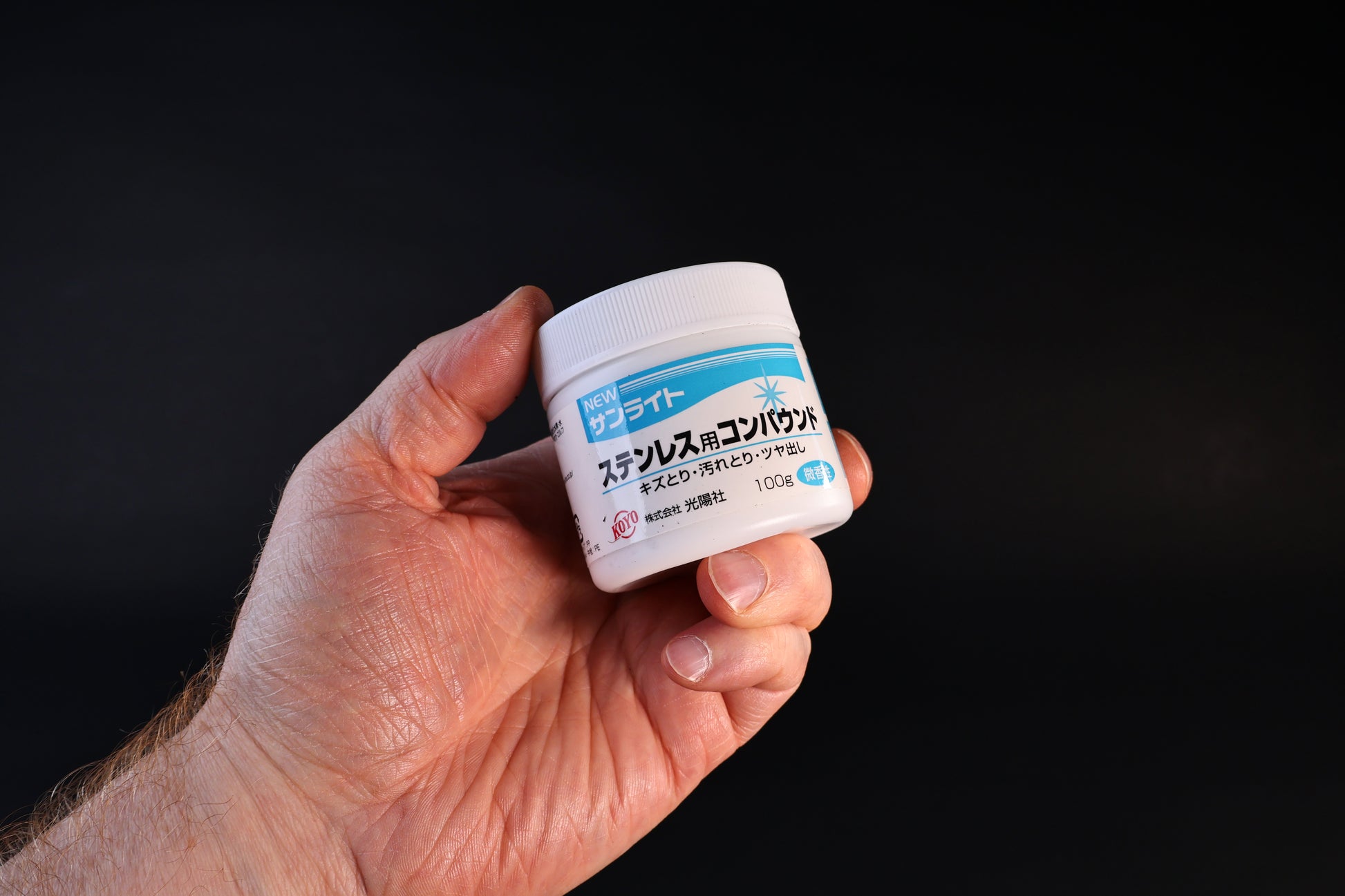 Koyo New Sunlite stainless steel polishing compound in a 100g white jar with blue label, held against a dark background