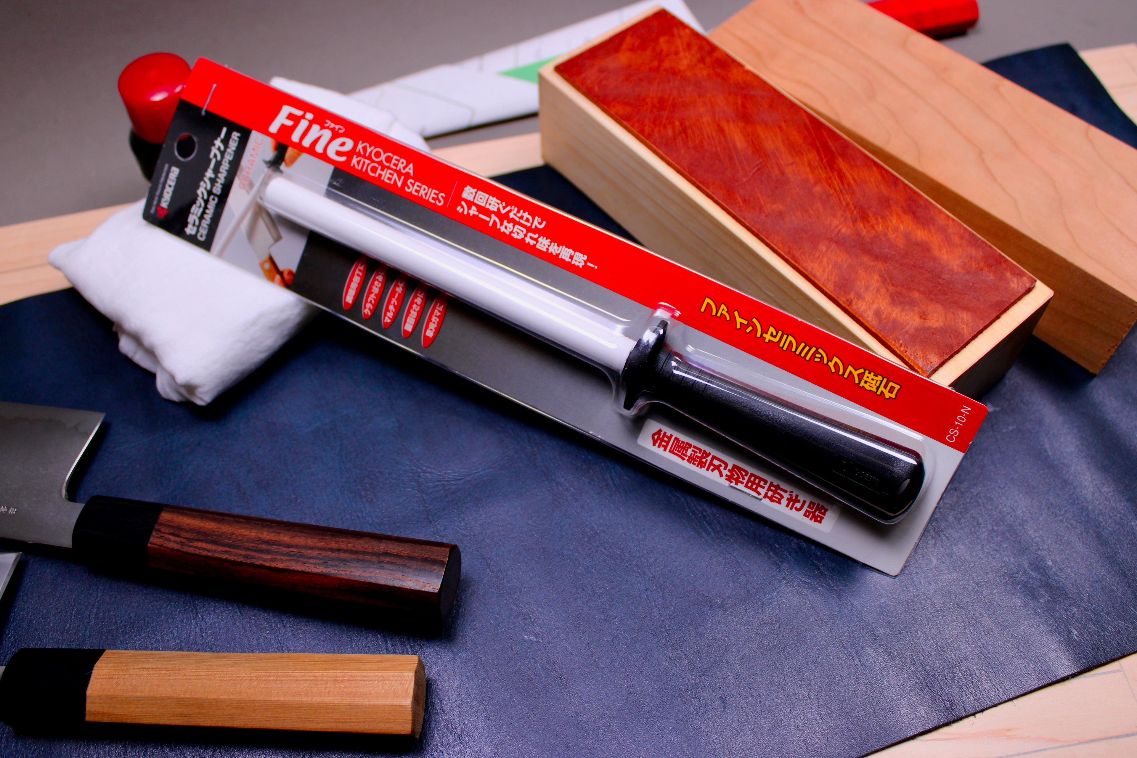 plastic black handled white ceramic honing rod in product packing with bright red and grey paper with hiragana writing surrounded by japanese knives leather strop hinoki wood 