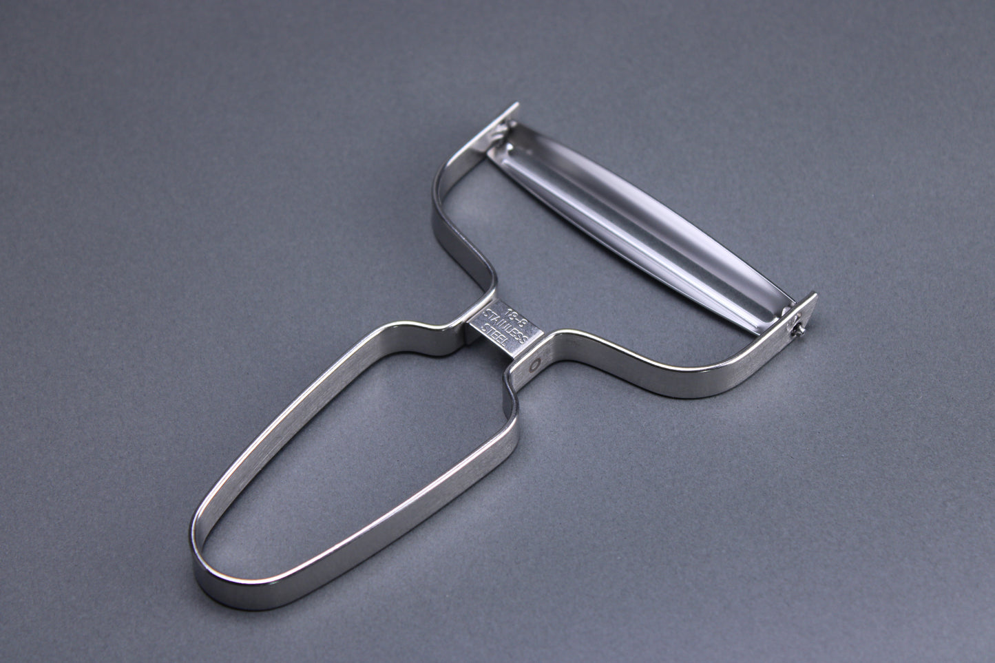 close up shot of metal vegetable peeler with curved angles and wide peeling surface imported from japan showing a curved blade and a grey background