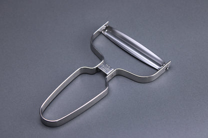 close up shot of metal vegetable peeler with curved angles and wide peeling surface imported from japan showing a curved blade and a grey background