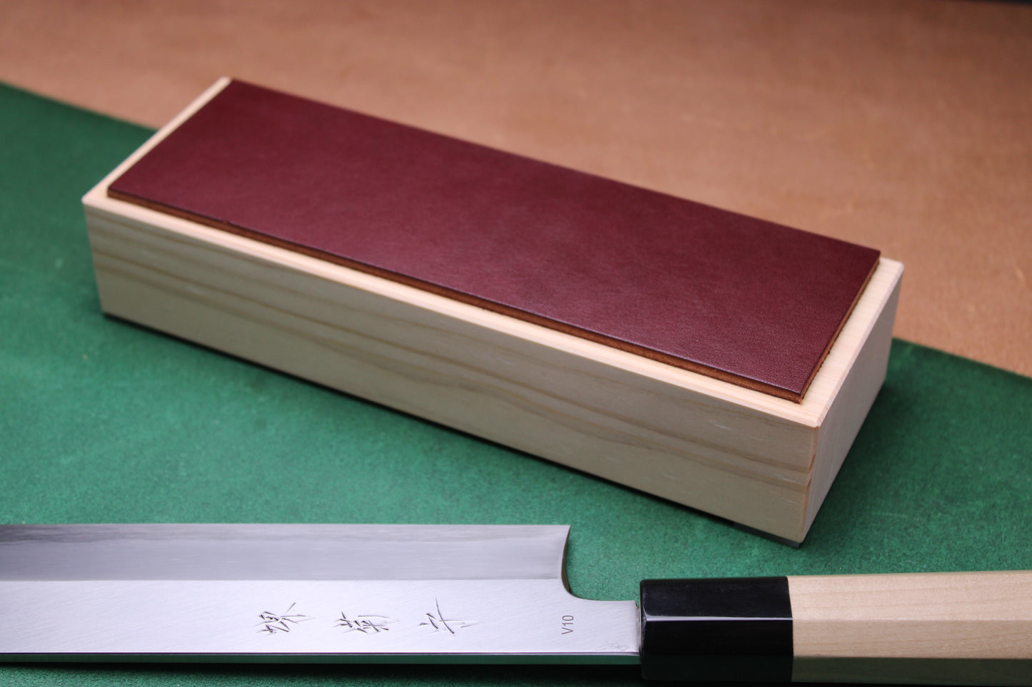 knife strop made of English bridle leather colored burgundy espresso brown attached to sturdy tall smooth hinoki wood base sakai kikumori japanese usuba vegetable knife foreground with background faded green and brown 
