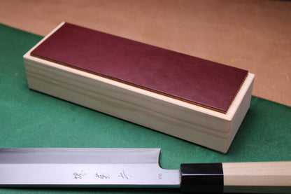 knife strop made of English bridle leather colored burgundy espresso brown attached to sturdy tall smooth hinoki wood base sakai kikumori japanese usuba vegetable knife foreground with background faded green and brown 