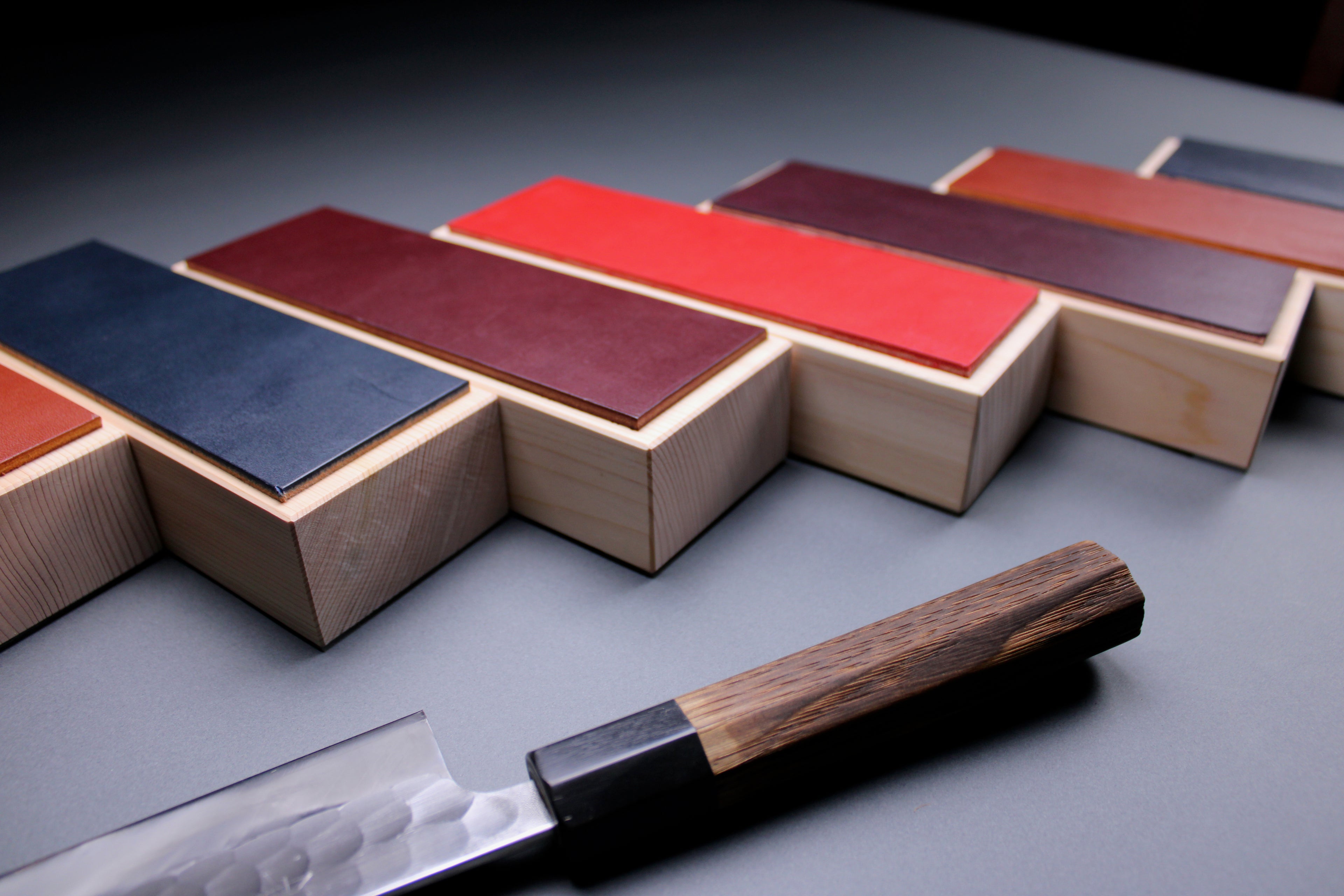 low close up shot of seven handmade leather knife strops and a yoshikane gyuto with a wood handle and a grey and black background