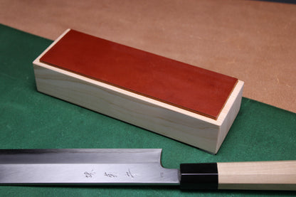 knife strop made of English bridle leather colored ochre tan brown attached to sturdy tall smooth hinoki wood base sakai kikumori japanese usuba vegetable knife foreground with background faded green and brown
