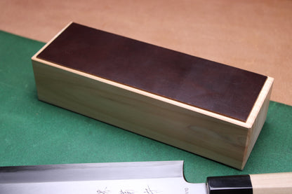 knife strop made of English bridle leather colored pecan deep brown attached to sturdy tall smooth hinoki wood base sakai kikumori japanese usuba vegetable knife foreground with background faded green and brown