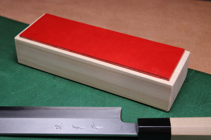 knife strop made of English bridle leather colored vermilion red attached to sturdy tall smooth hinoki wood base sakai kikumori japanese usuba vegetable knife foreground with background faded green and brown