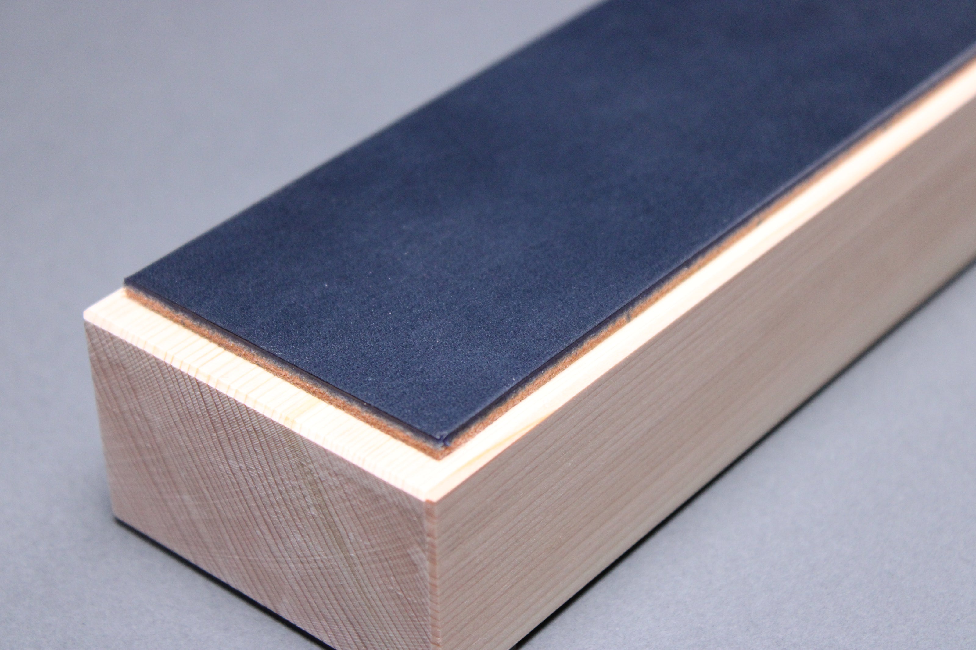 textured surface of midnight blue leather perfect for knife stropping atop sturdy tall hinoki wood base adequate height for knuckle clearance with grey background closeup shot