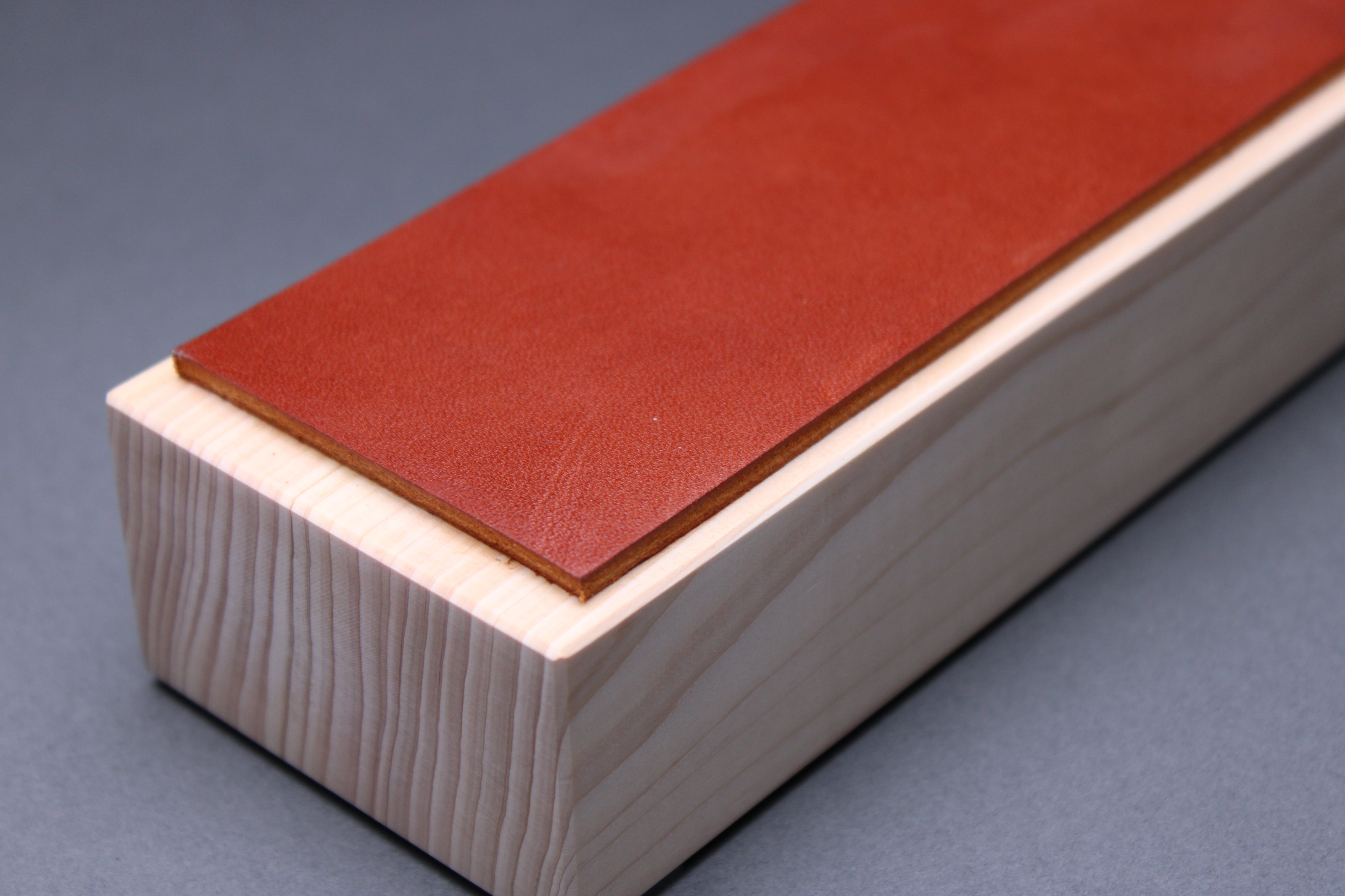  textured surface of ochre tan brown leather perfect for knife stropping atop sturdy tall hinoki wood base adequate height for knuckle clearance with grey background closeup shot