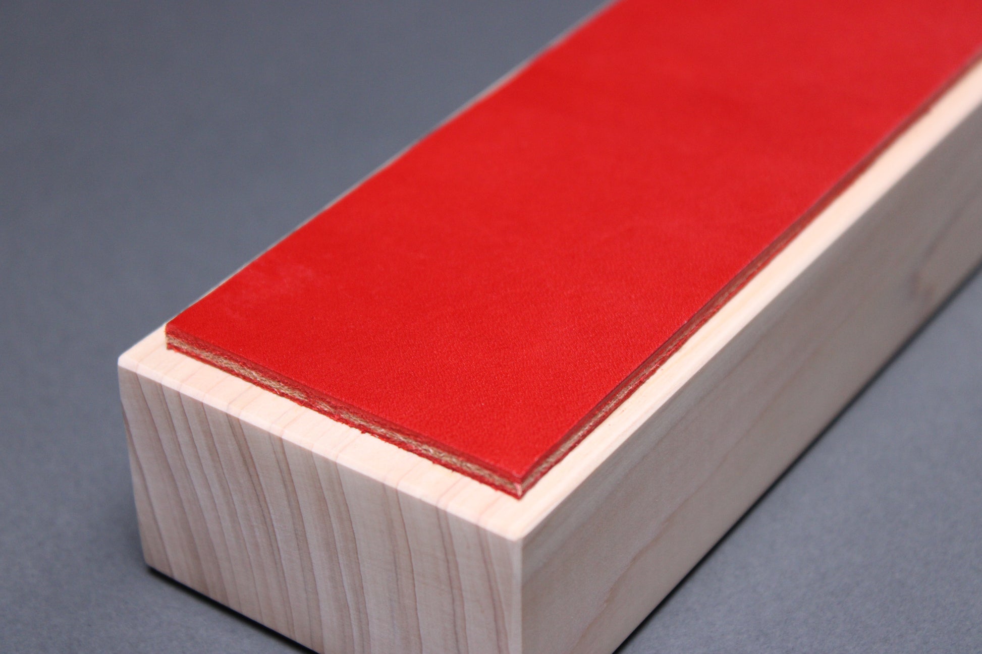 textured surface of vermilion red leather perfect for knife stropping atop sturdy tall hinoki wood base adequate height for knuckle clearance with grey background closeup shot