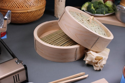  kitchen scene handmade bamboo and hinoki chinese steamer in foreground surrounded by portable stove hinoki cutting board ceramic plate of broccoli green cabbage scallions cooking chopsticks steam cloth 