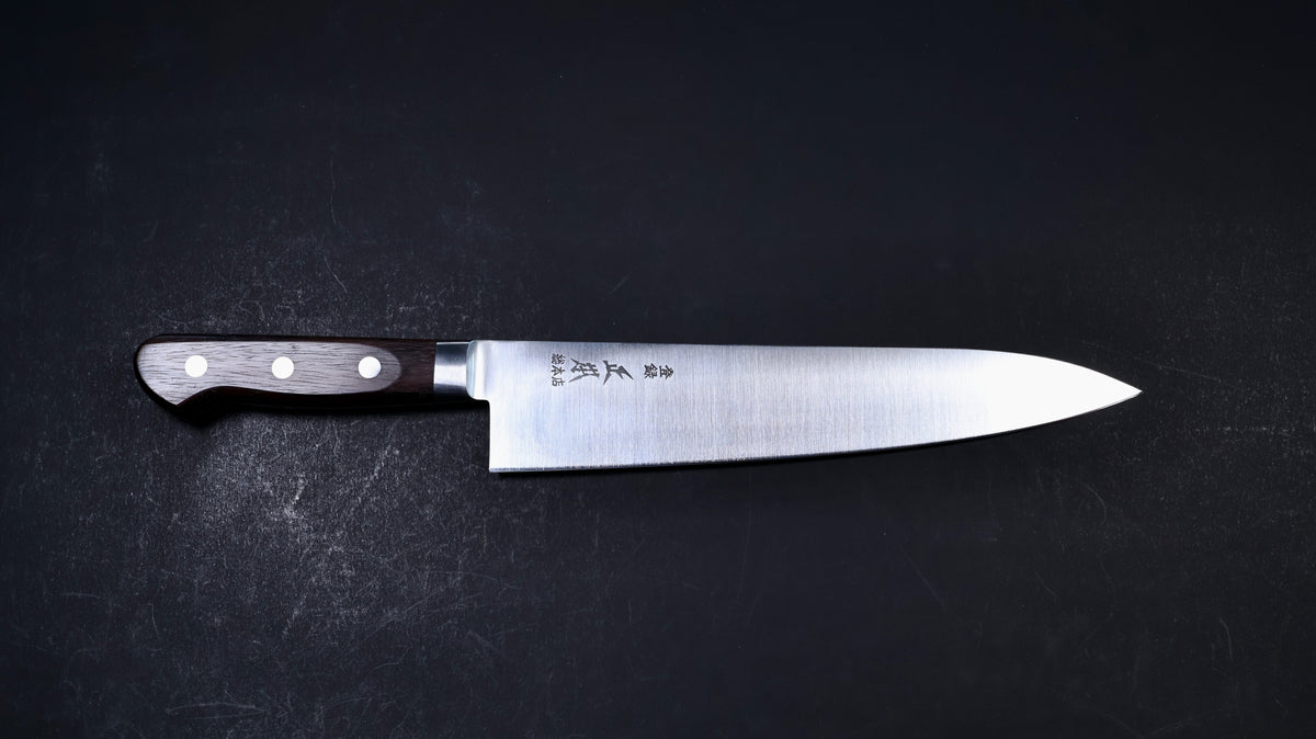 A Japanese chef's knife with a satin-finished silver steel blade and a dark brown wooden handle, displayed horizontally on a dark textured background. The blade features engraved Japanese characters and the brand name 'Yasuhiro'.