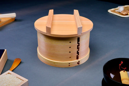 round natural wooden food steamer two handled lid atop body sitting on gray surface with japanese kitchenware in background  
