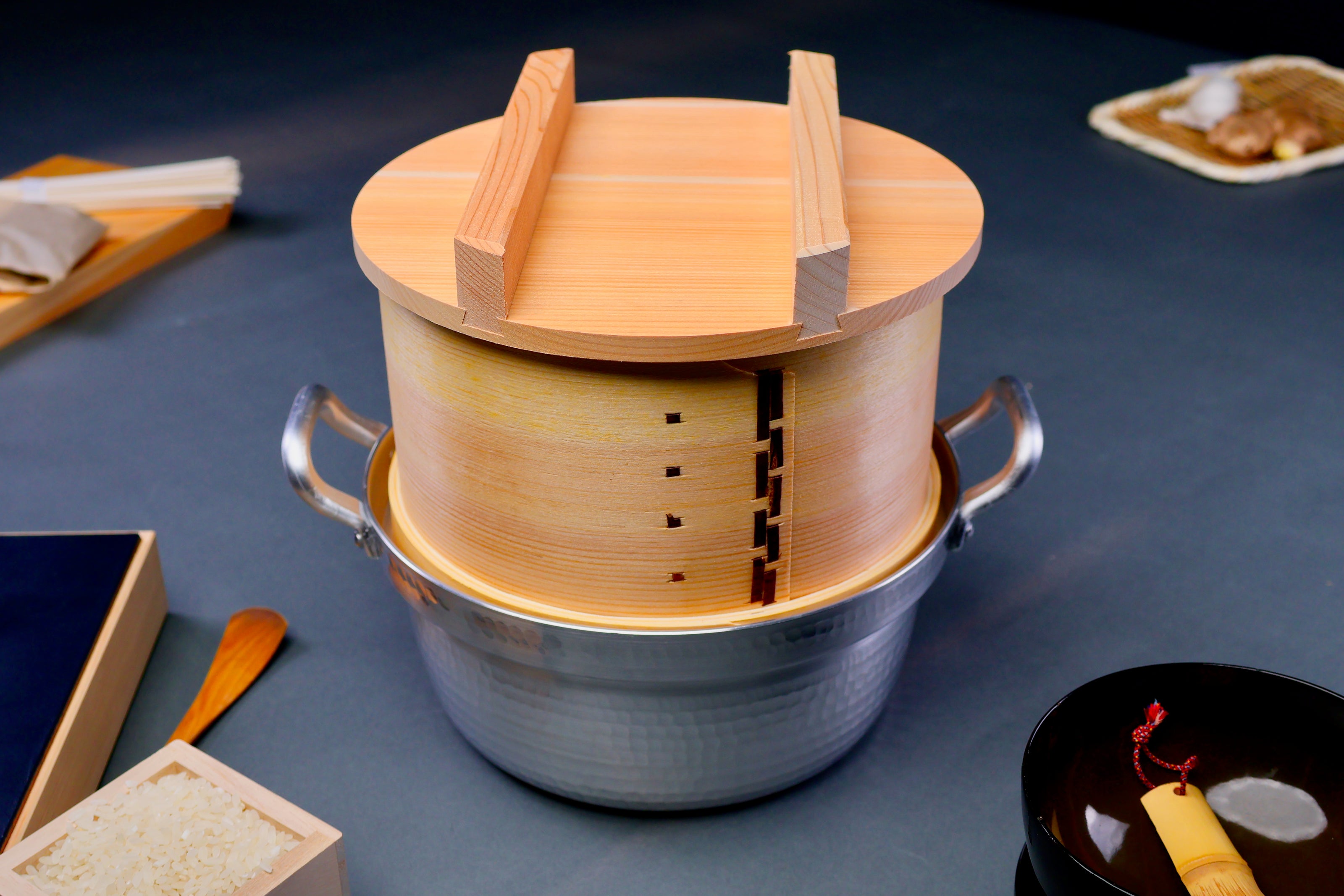 round natural wooden food steamer two handled lid atop body sitting on interior ledge of hammered aluminum two handled pot in grey background  