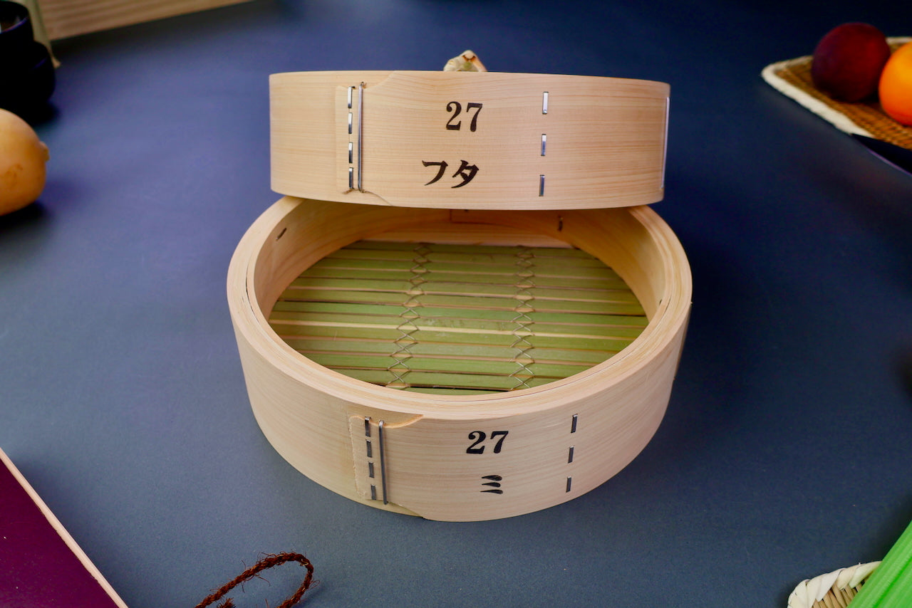 thick wood round bamboo steamer set woven top with loop natural wood tone with printed hiragana surrounded by japanese kitchenware blue gray background