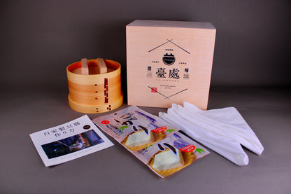 round wooden tofu steamer with two handled lid next to pale wood gift box with and two white towels plastic packaging and gift set pamphlet with hiragana writing on grey surface