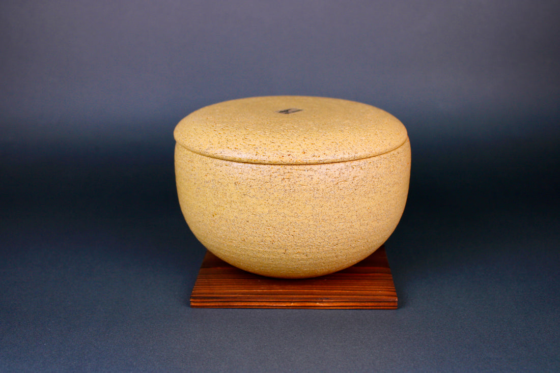 short round yellowish ceramic ohitsu sitting atop tan square wood trivet in dark grey background