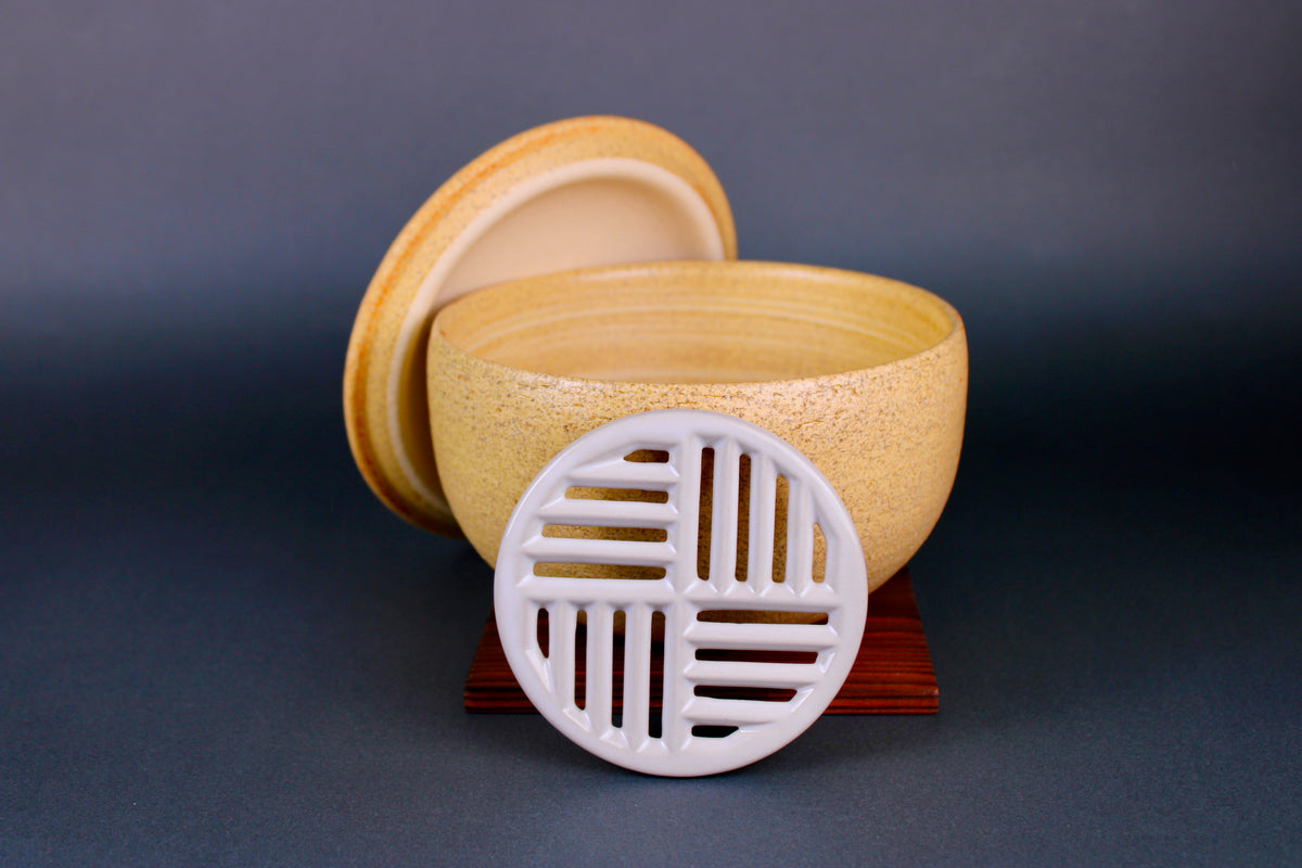 short round yellowish ceramic bowl with white grate leaning against main body atop tan wood square trivet lid in background surrounding grey backdrop