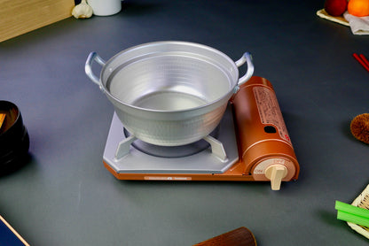 two handled stepped aluminum silvery pot with shiny hammered surface sits atop portable silver tan butane stove on a dark grey backdrop 
