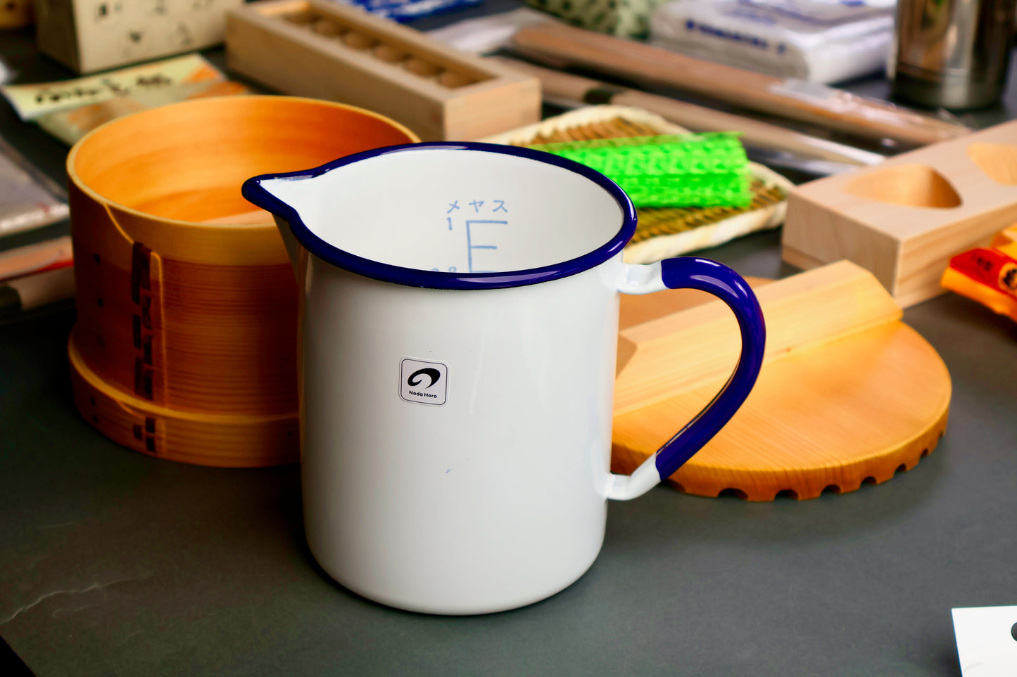 White enamel one liter measuring cup with hiragana noda horo label with blue rim and handle on a cluttered surface