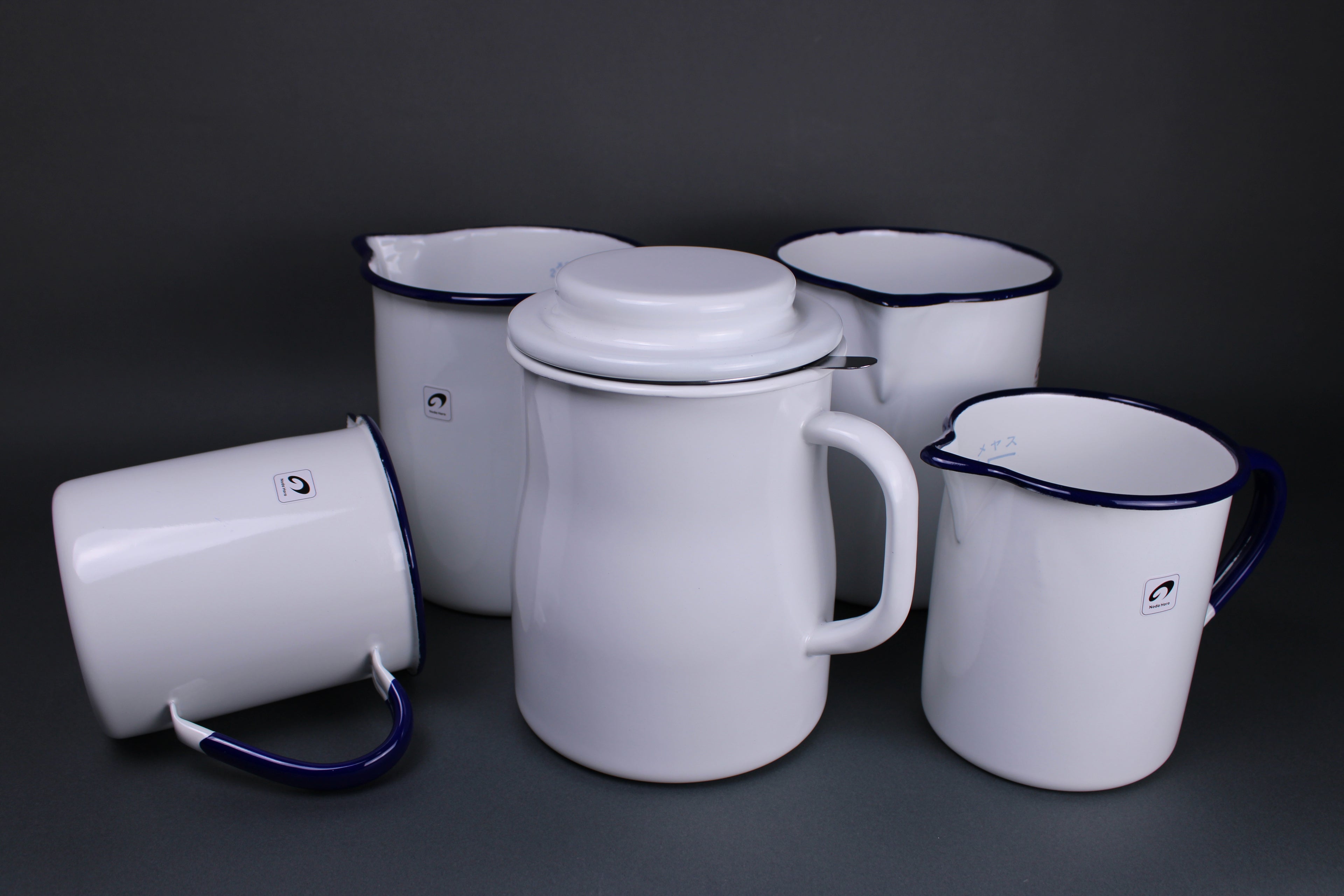 front facing image of noda horo four measuring cups and one storage pot for kitchen oil one cup is laying on its side all are glazed a creamy white with curved rims and stout large handles the products are japanese kitchenware 