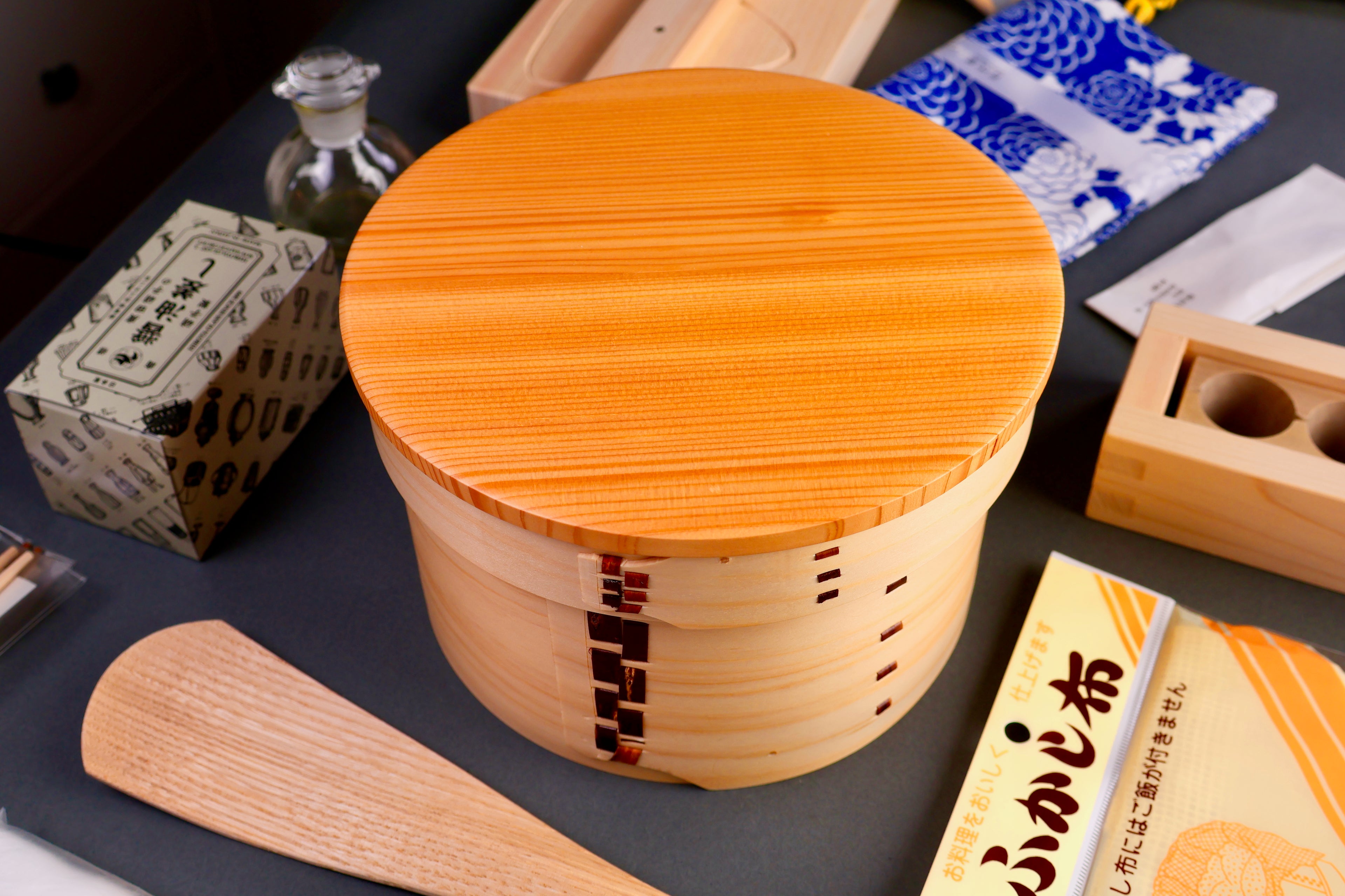 round hinoki wood container with lid surrounded by japanese kitchenware in dark grey background