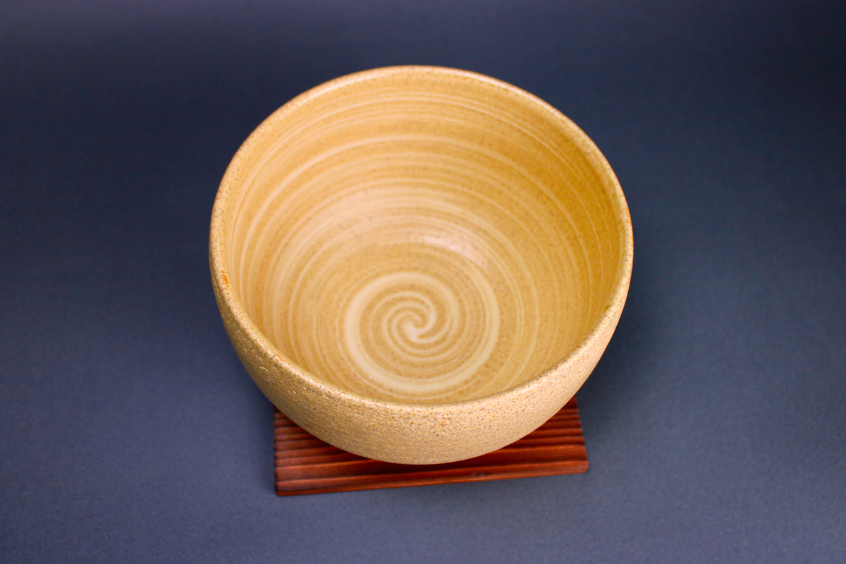 short round yellowish ceramic bowl sitting atop tan wood square trivet in grey background