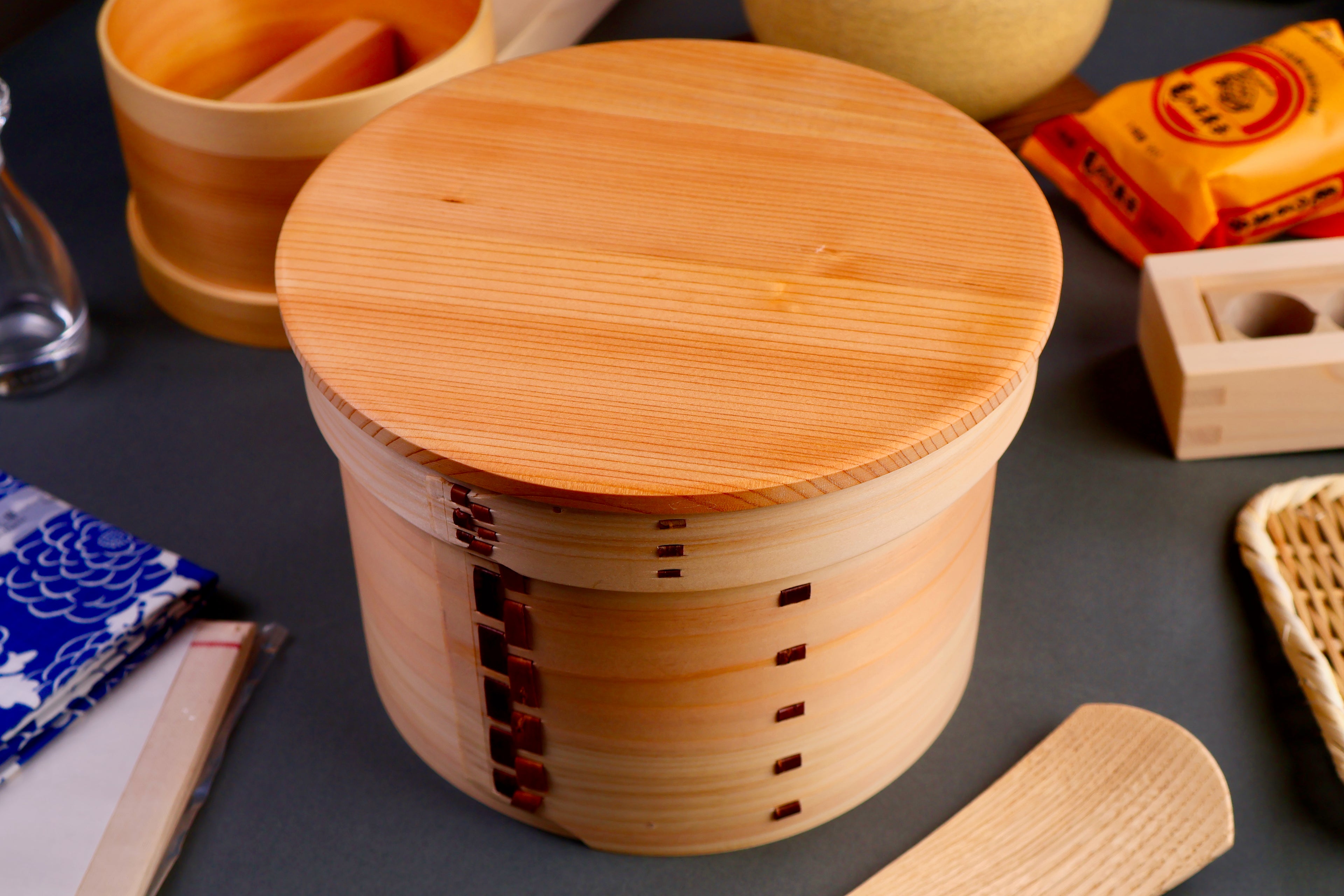 round hinoki wood container with lid surrounded by japanese kitchenware in dark grey background