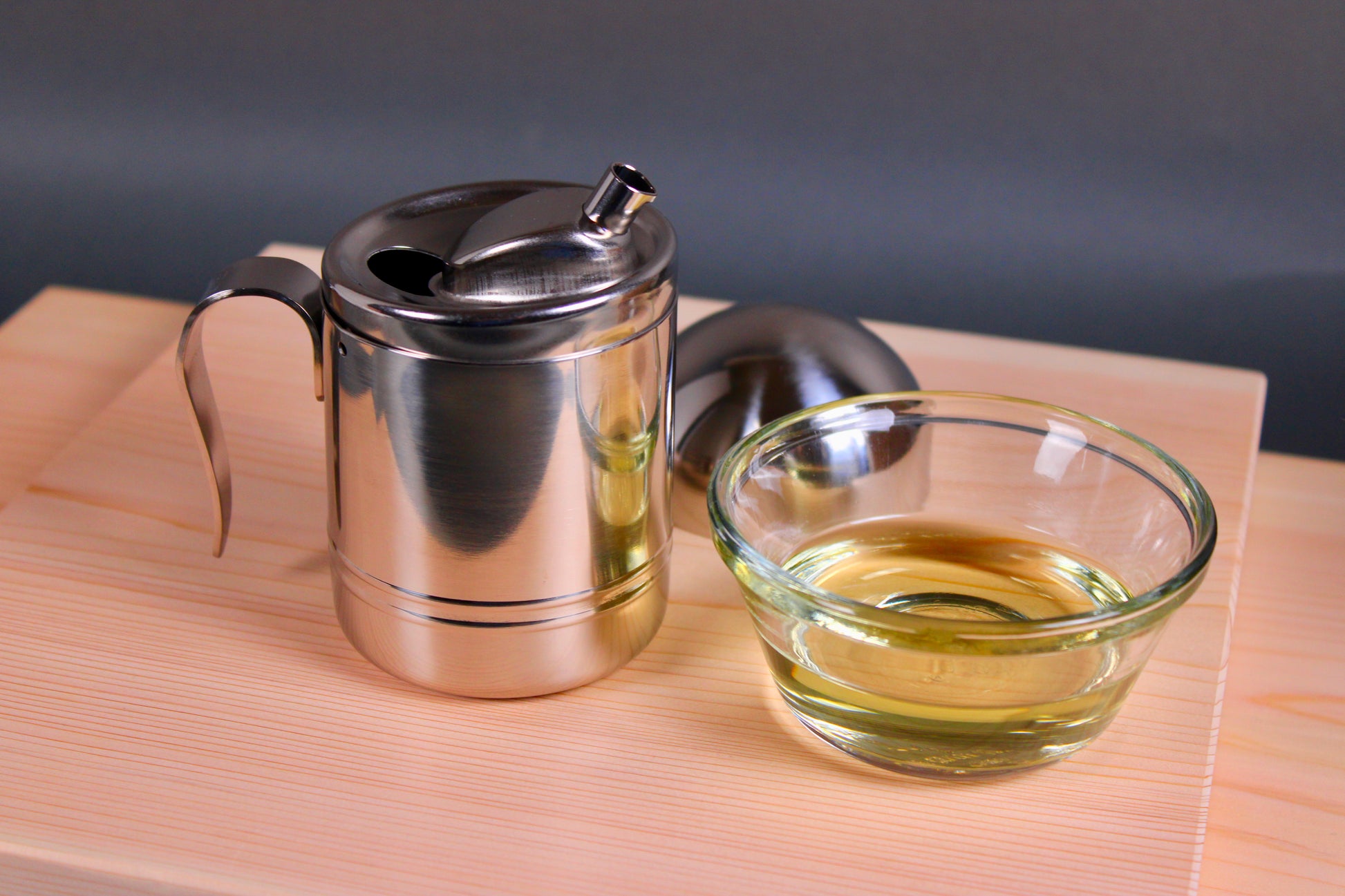 stainless steel round oil cruet with curved handle narrow spout domed lid beside container beside clear glass bowl filled with glossy oil sitting atop two hinoki cutting boards 