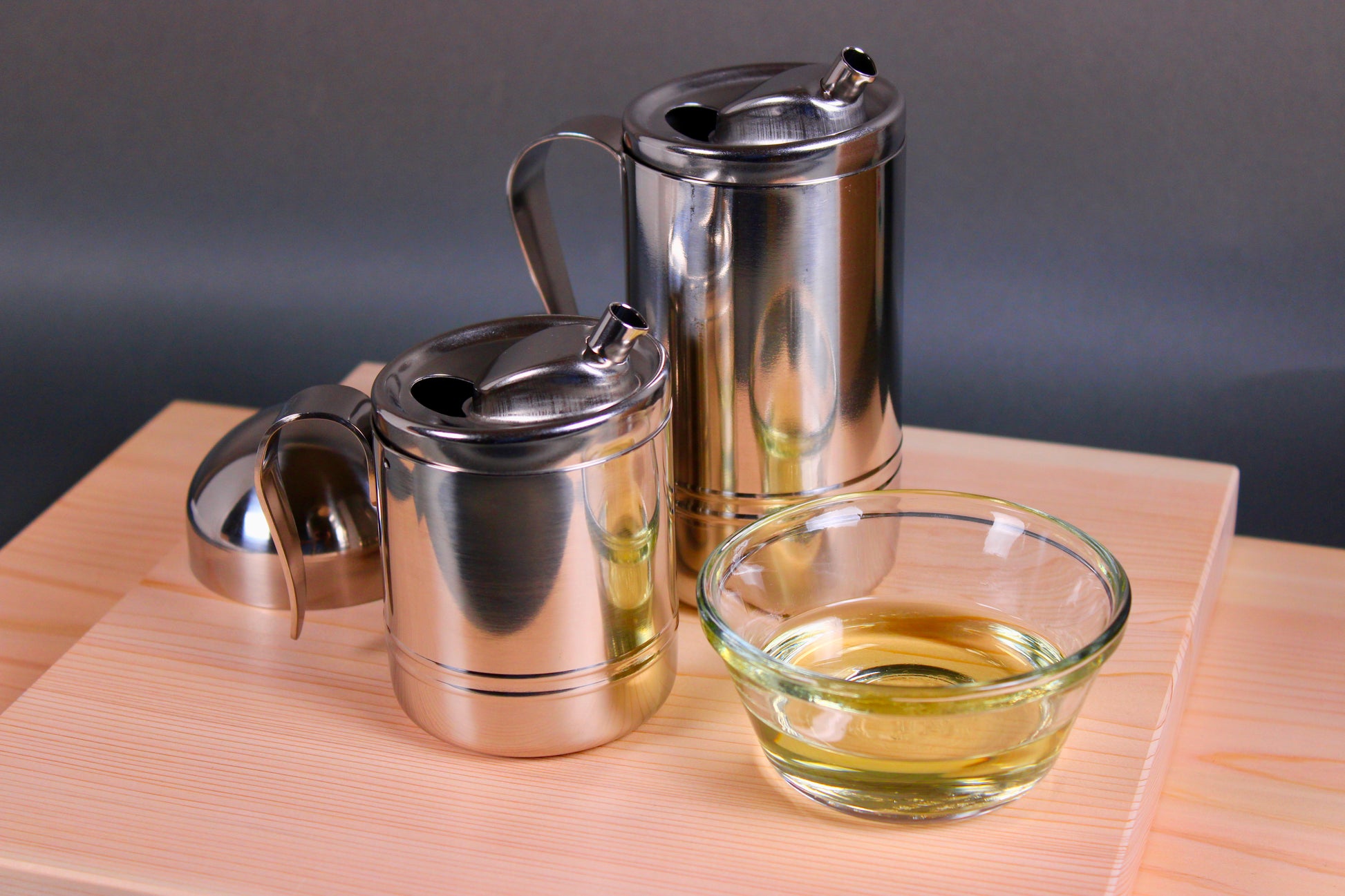 two stainless steel oil containers for japanese cooking next to glass bowl with oil atop wood cutting boards