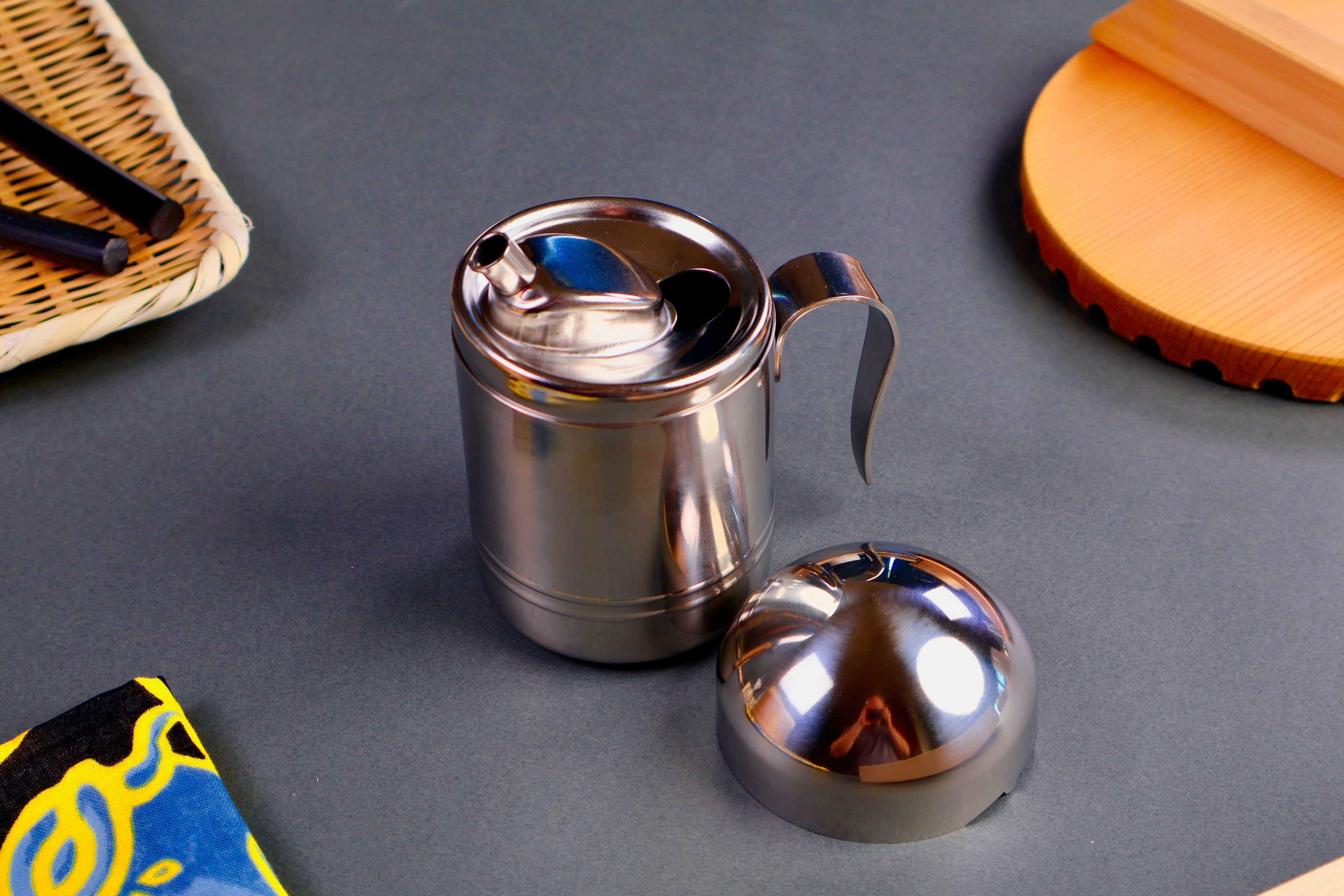 Stainless steel  round oil dispenser with curved handle metal spout surrounded by japanese kitchenware domed lid  on a gray surface